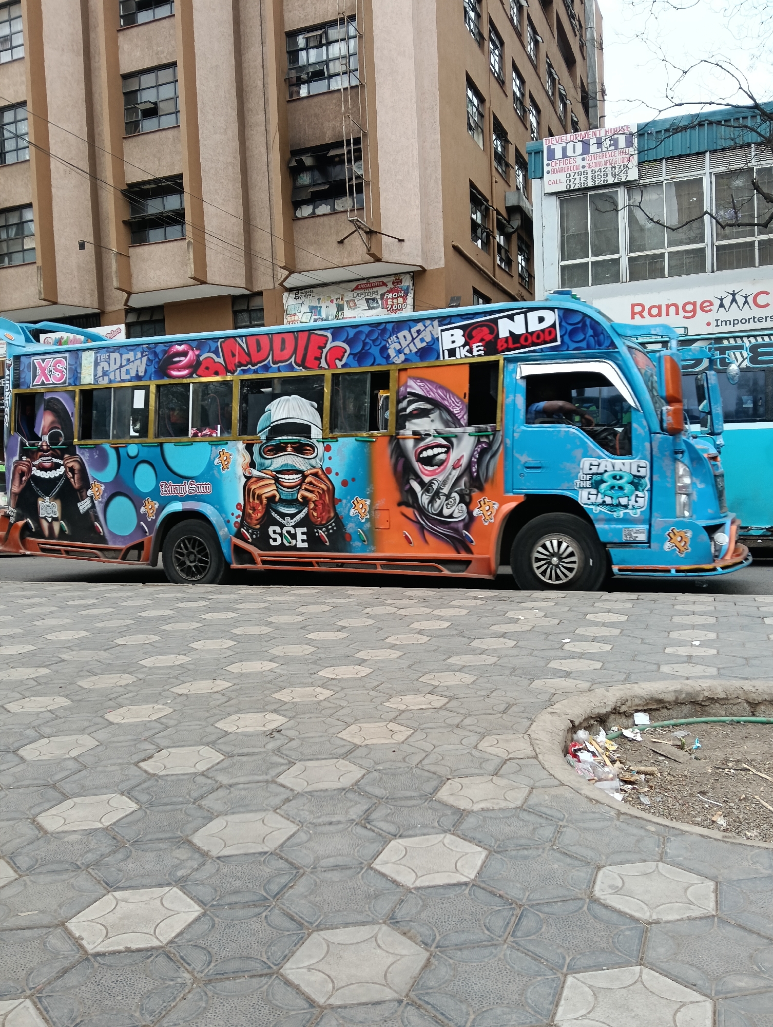 The image is a street-level, medium shot of a vibrant, graffiti-covered matatu (public transport minibus) parked on a street in Nairobi, Kenya. The matatu is painted primarily in bright blue with extensive artwork featuring stylized portraits of individuals with exaggerated features, jewelry, and clothing. Text adorns the vehicle, including "XS", "THE CREW", "BADDIEs", "BOND LIKE BLOOD", and "GANG OF THE 8 GANG". The matatu has tinted windows and chrome accents.
The background features a multi-story building with a beige facade and numerous dark-framed windows. Signs are visible on the building, including one that reads "DEVELOPMENT HOUSE" with a list of services such as "OFFICES", "CONFERENCE HALL", "BOARDROOM", and "RESEARCH AREA & LIBRARY". Another sign to the right indicates "Range importers".
The foreground shows a patterned pavement made of interlocking hexagonal and rectangular tiles in shades of gray and white. To the right of the pavement, there is a small area of disheveled ground with scattered trash, including plastic bags, paper, and debris, adjacent to a green hose. The sky is overcast and appears white, suggesting a cloudy day. The overall scene depicts a typical urban street environment in Nairobi, characterized by the distinctive artistic style of its public transportation.
