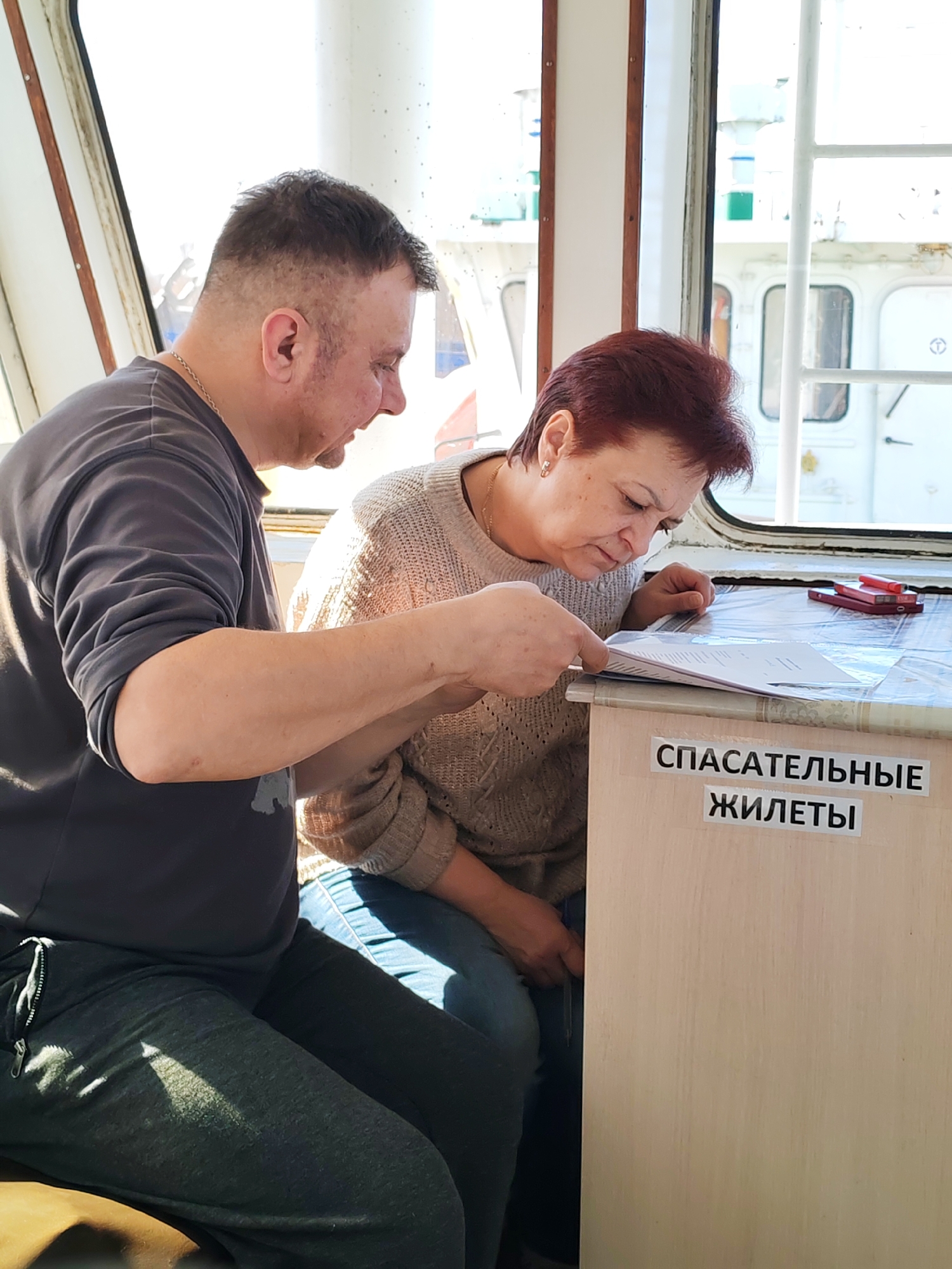 An indoor, brightly lit photograph captures two individuals, a man and a woman, seated and focused on documents within what appears to be the cabin of a marine vessel, such as a boat or ferry. This is suggested by the windows with nautical-style frames and the view of a large white structure outside, possibly another part of the ship or a nearby vessel.
On the left, a man with short, dark, buzzed hair wears a dark grey short-sleeved t-shirt over a darker long-sleeved shirt and dark grey sweatpants, along with a thin chain around his neck. He leans forward, intently looking down at the papers held by the woman, with his right arm extended towards them as if pointing.
On the right, a woman with short, reddish-brown hair is dressed in a light beige knitted sweater and blue jeans. She is deeply engrossed in studying the papers, holding them with both hands on a light wooden table or cabinet. A red smartphone and a red pen are also visible on the table surface next to the documents.
The bright natural light streaming through the windows indicates it is daytime, likely a clear day. Both individuals appear serious and concentrated on the task at hand, suggesting they are reviewing or discussing the contents of the papers.
A prominent detail is the text displayed on the side of the wooden table/cabinet. Two black-on-white labels read "СПАСАТЕЛЬНЫЕ" (SPASATEL'NYE) and "ЖИЛЕТЫ" (ZHILETY), which collectively translate from Russian to "LIFE VESTS" or "RESCUE VESTS," further confirming the marine setting and the cabinet's likely purpose for storing safety equipment. The overall atmosphere is one of focused attention within a functional environment.