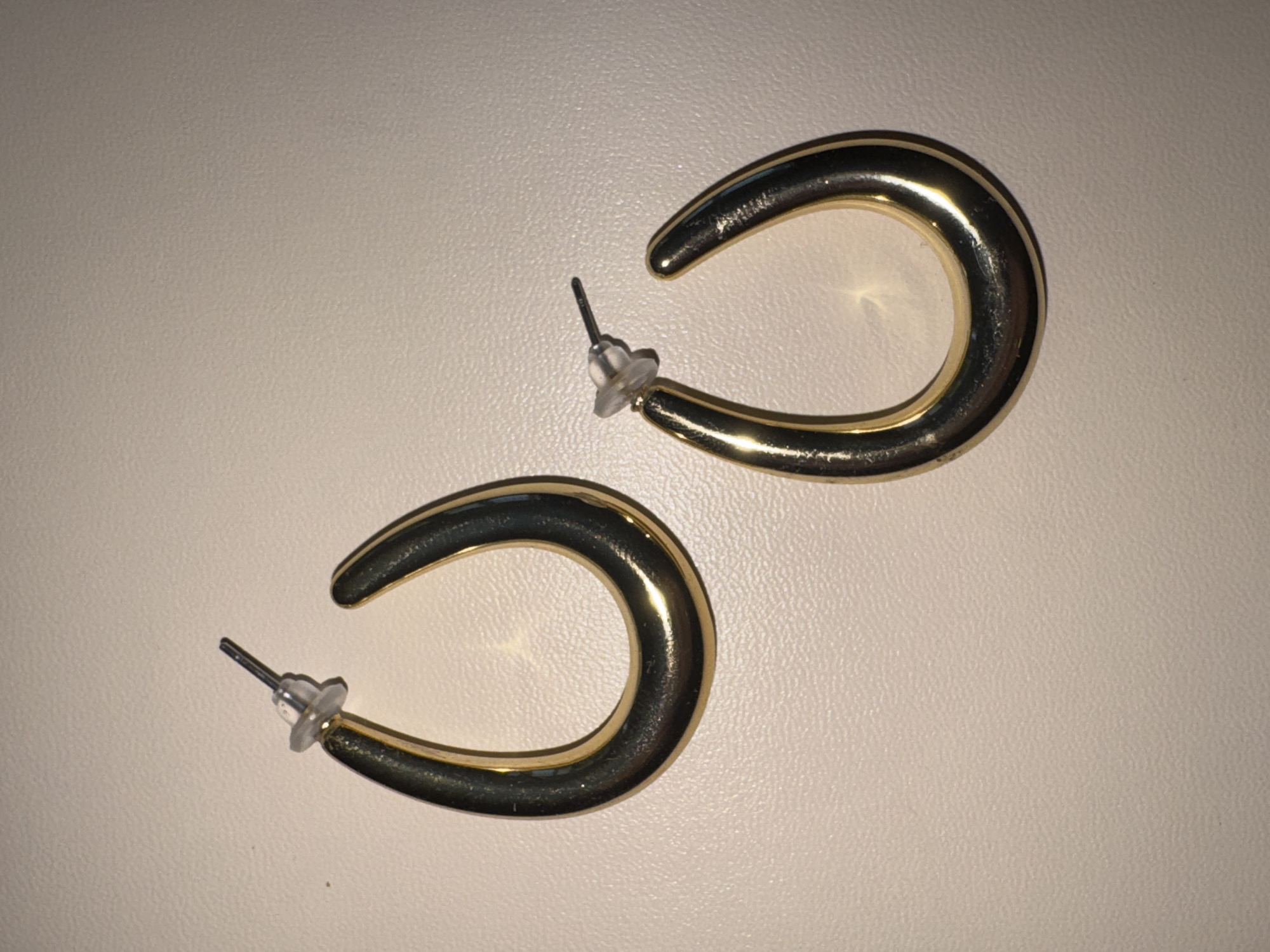 The image shows a pair of gold-colored hoop earrings lying on a light-colored, textured surface. The earrings are shaped like flattened ovals or teardrops. One earring is positioned above and to the right of the other. Each earring has a post and a clear plastic backing, indicating they are designed for pierced ears. The surface appears to be a tabletop or similar flat surface, possibly indoors. There is no visible text, and the lighting suggests it is daytime, though no specific location cues are present beyond the general indoor setting. The focus is solely on the jewelry.