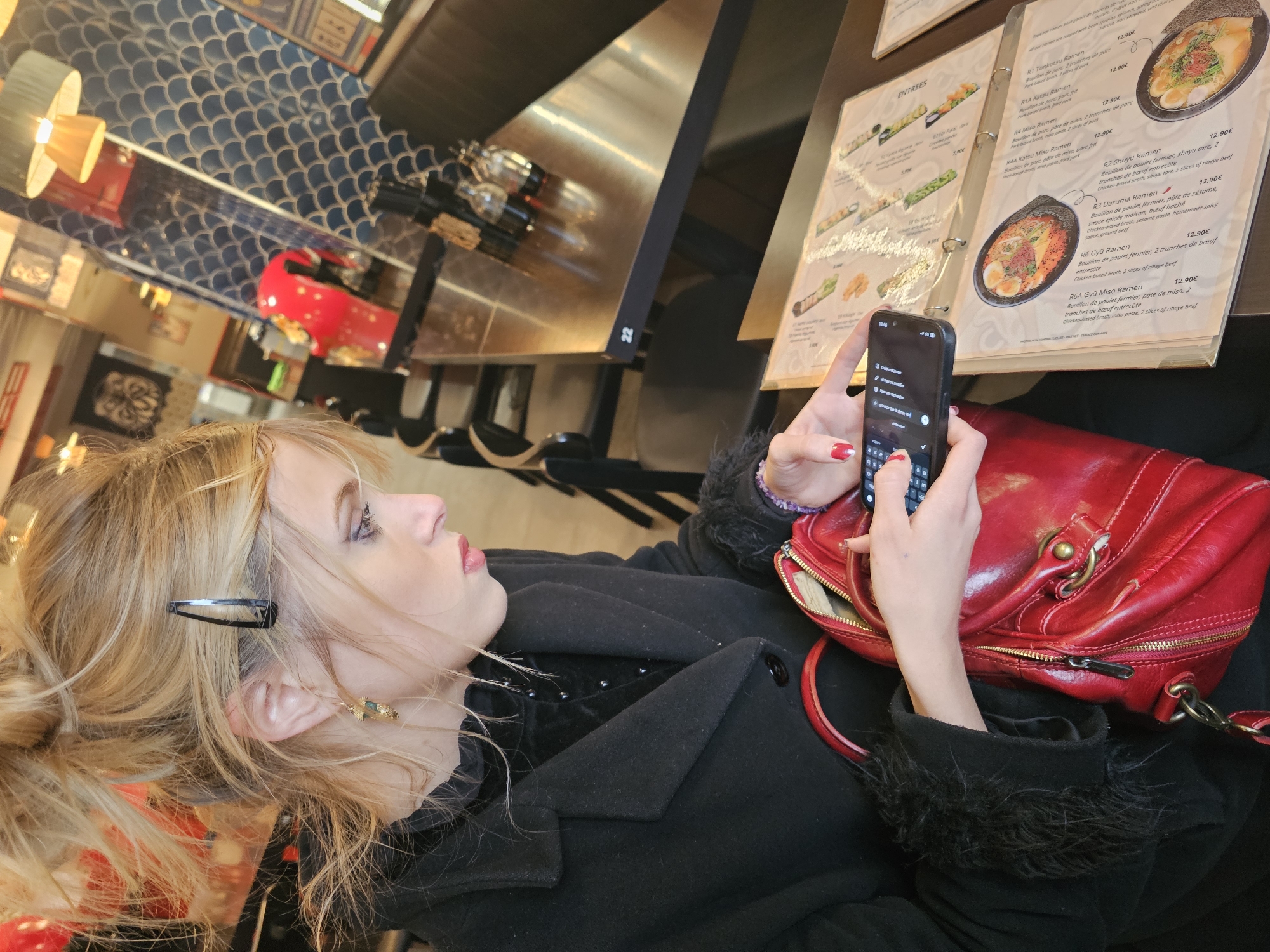 A fair-skinned woman with blonde hair is seated indoors, likely at a restaurant, and using her black smartphone. She is wearing a black coat with a fur-like trim. Next to her is a red handbag. The focus of her attention is the phone's screen. Beside her are restaurant menus listing various ramen dishes, with prices in euros. The background features a bar area with bottles and patterned wall tiling. The location is implied to be in Paris, France, based on the language on the menu.