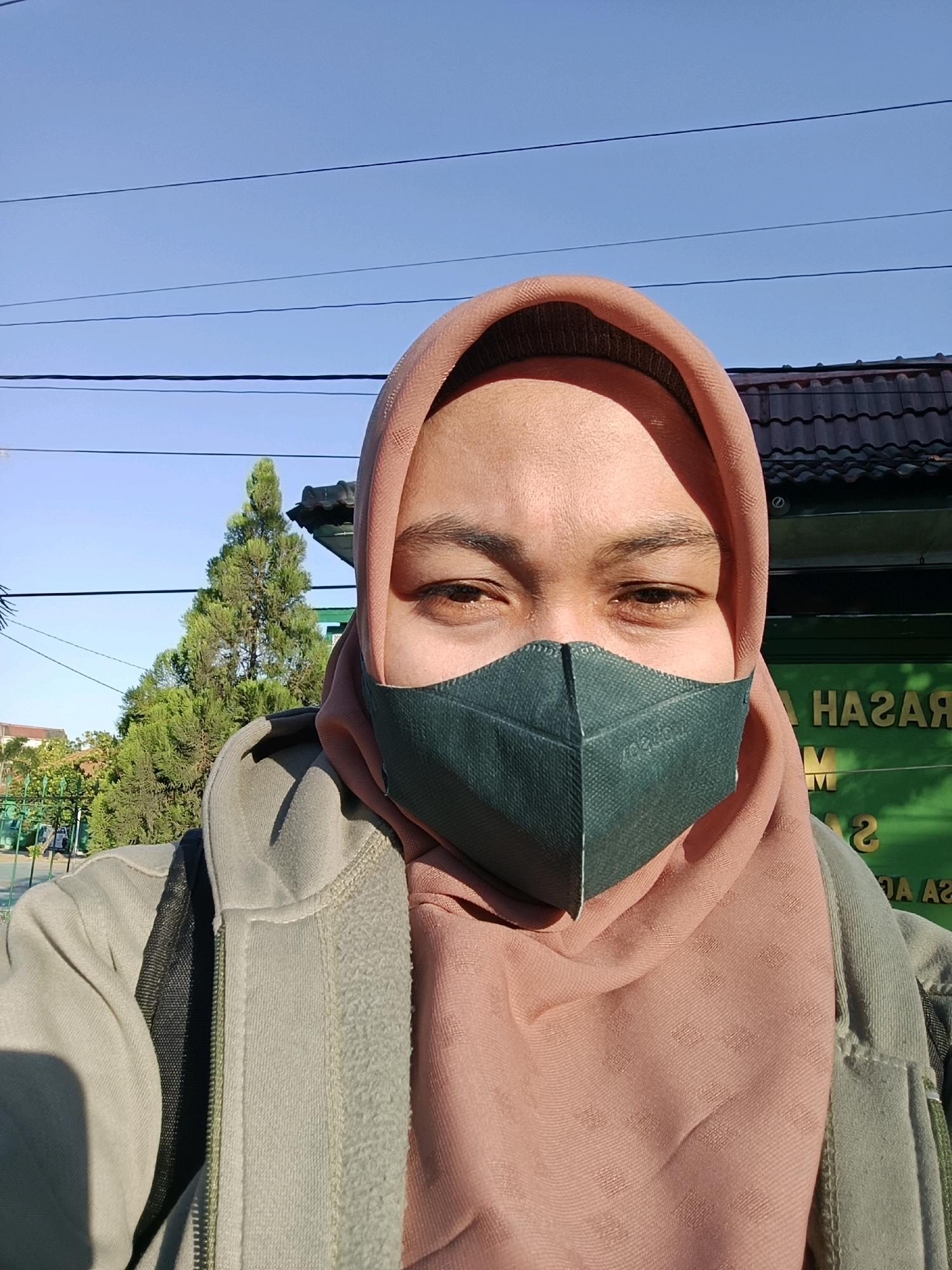 A close-up, selfie-style photograph captures a person wearing a light brown hijab and a dark green face mask. The individual's visible face, from the eyebrows down, is illuminated by sunlight, creating a shadow on their forehead. They are wearing a light olive green zip-up hooded jacket over a pink garment, possibly another layer of clothing or a scarf. The background is blurred but shows a bright blue sky with thin power lines, a tall evergreen tree, and a building with a dark, tiled roof and green walls. Faint text, possibly signage, is visible on the green wall. The overall setting appears to be outdoors, in a sunny environment within the area of Gunung Sekar, Indonesia.