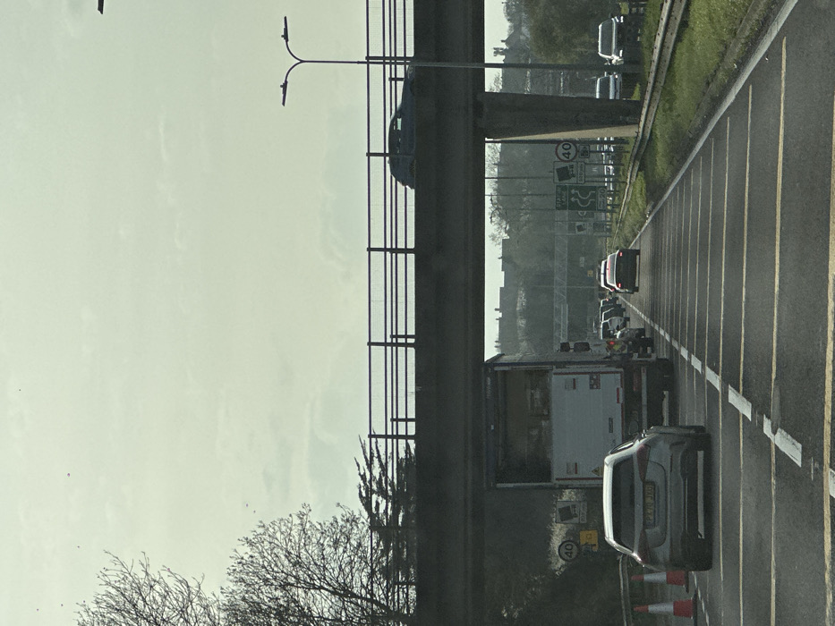 The image displays a multi-lane road experiencing traffic congestion under a concrete overpass in Basildon, United Kingdom. This location is identified as the A127 (Southend Arterial Road) due to visible directional signs for "M25" and "A127" on an overhead gantry further down the road, coupled with a "40" mph speed limit.
The perspective is from within a vehicle, facing eastbound. In the foreground, two orange traffic cones are positioned on the left shoulder. A silver Mercedes-Benz A-Class hatchback (UK registration PX70 JXG) is stationary or slow-moving in the left lane. In the adjacent lane, a large white box truck with rear red and white chevron markings is situated directly beneath the overpass, also stationary or slow-moving. A queue of additional vehicles, including a dark sedan with illuminated taillights, extends into the distance, indicating traffic.
The concrete overpass spans the road, featuring a black metal railing on its upper edge. A dark SUV or similar vehicle is observed moving across the overpass from left to right. A tall black streetlight with a double-headed luminaire is positioned on the right side of the overpass. On the left side of the road, bare deciduous trees and evergreen shrubs are visible. Road signs include a circular "40" mph speed limit sign and, further ahead, an overhead gantry displaying directional signs for "M25," "A127," and a congestion warning. The road surface features distinctive yellow horizontal rumble strips and white lane markings. The right side of the carriageway has a grassy verge and a metal crash barrier. The sky is overcast and pale grey. Distant urban structures are faintly visible on the horizon.