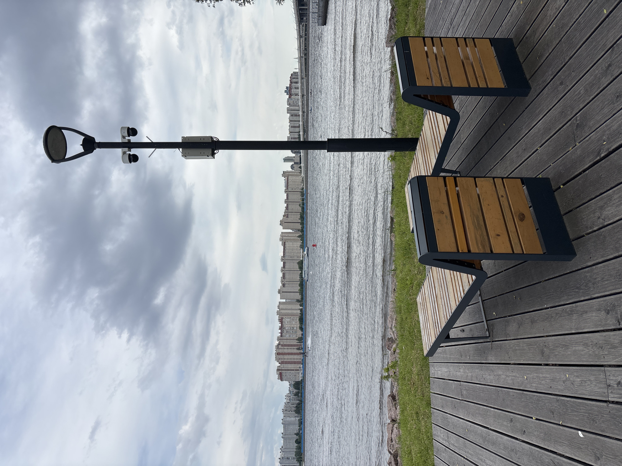 The image captures a serene waterside promenade in Saint Petersburg, Russia, under an overcast sky during the daytime. The foreground features a dark wooden plank deck, along which two modern benches are positioned. These benches have slatted wooden seats and backrests, supported by dark metal frames.
To the left, a tall, black lamppost stands prominently, equipped with a contemporary street light fixture at the top. Attached to the pole are two white security cameras and a grey utility box, suggesting urban infrastructure for surveillance or smart city functions.
Beyond the deck, a narrow strip of green grass and scattered rocks borders a wide body of water, likely a river or bay. The water's surface shows subtle ripples. Across the water, a long stretch of urban landscape unfolds, dominated by numerous multi-story residential buildings lining the distant shore. In one of the closer views, a white boat and a small red buoy are visible on the water. A large bridge structure can also be seen in the far distance, spanning the water.
The sky is filled with a dense layer of grey clouds, indicating cloudy weather. The scene is devoid of people, conveying a quiet, contemplative atmosphere. No visible text is clearly legible in the image.
