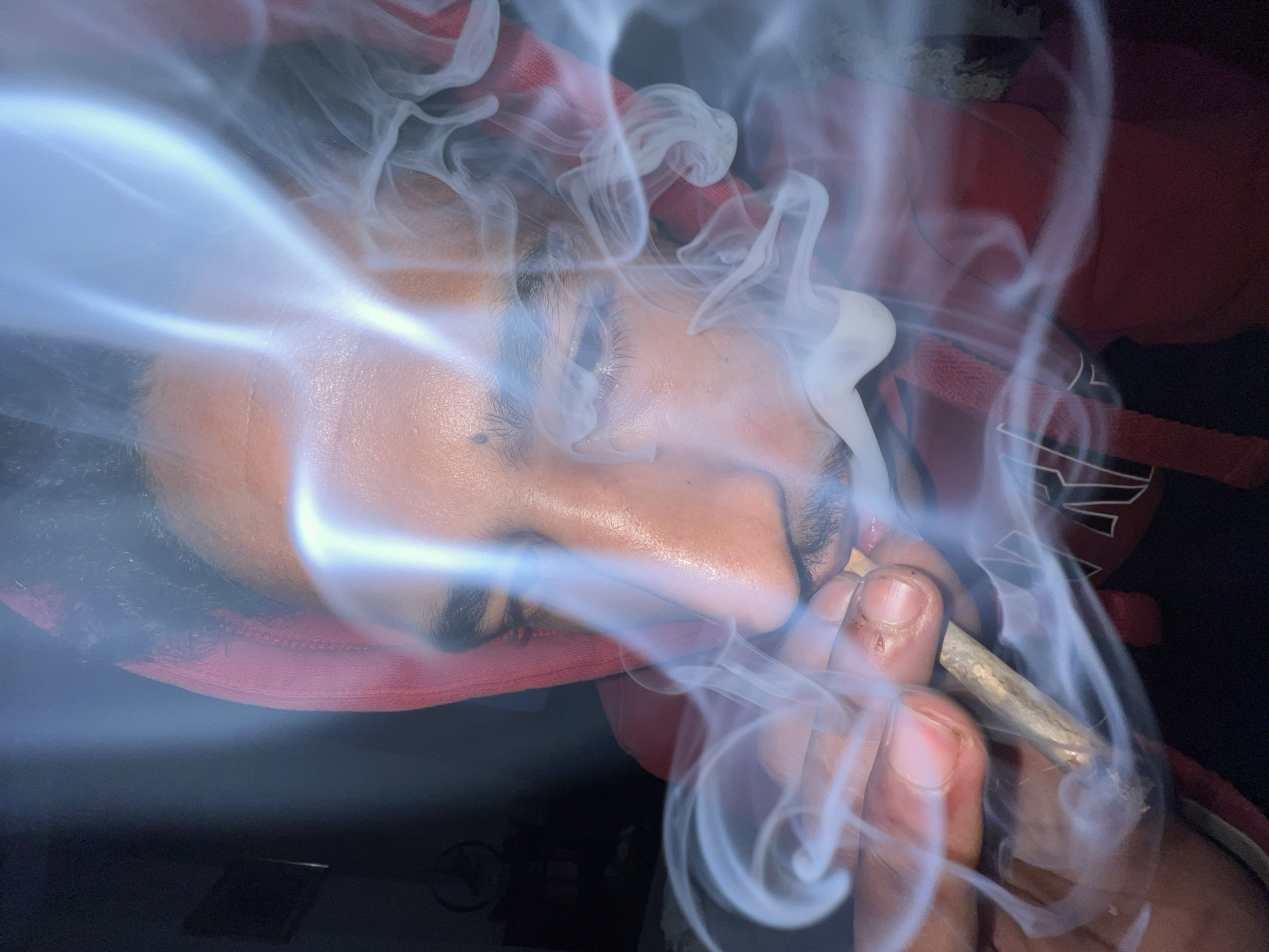 The image depicts a close-up, low-light portrait of a single individual, consistent with a male of North African ethnicity, engaged in smoking a hand-rolled cigarette indoors in what is understood to be a private setting within Tanger, Morocco. The subject is centrally positioned, facing slightly left of the viewer, with their head angled downward. They wear a red hooded garment with the hood up, partially obscuring dark hair. A faint white graphic is visible on the front right side of the red garment. The person's right hand holds the lit, hand-rolled cigarette between the thumb and index finger, bringing it to the mouth. Dense, light-colored smoke emanates from the cigarette and swirls prominently in the foreground and around the individual's face. Visible facial features include the right eye, nose, mustache, and a portion of the mouth. The background is dark and out of focus, revealing a faint framed object on the upper left wall and, below it, a circular decorative item featuring a star-like motif. The scene is illuminated by an unseen light source that highlights the smoke and parts of the subject's face.