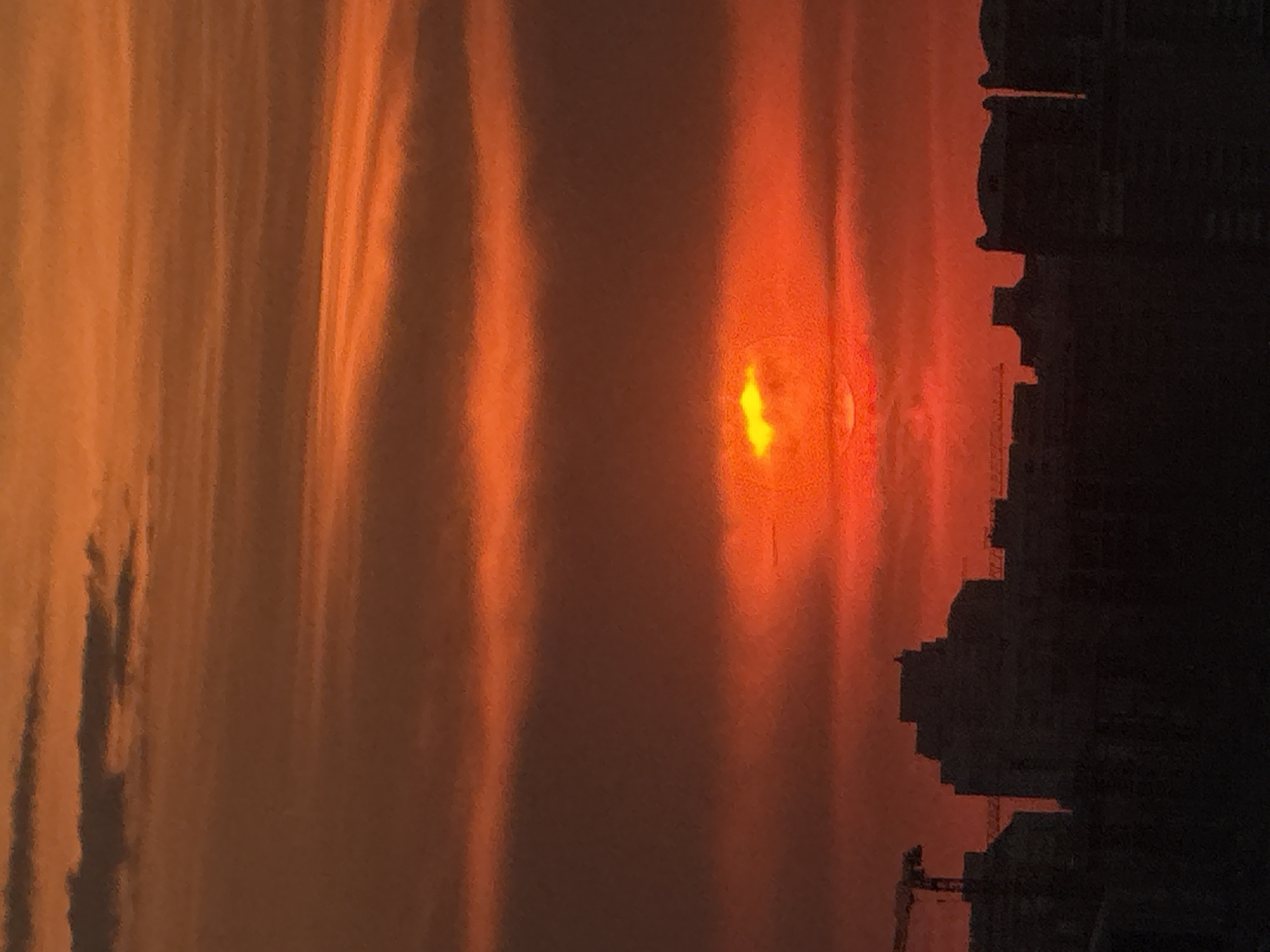 The image shows a sunset in a city, likely Moscow, Russia based on the architecture of the buildings in silhouette on the right. The sun is a bright orange orb with some visible detail around it, positioned in the center of the frame. The sky is filled with streaks of orange and yellow, creating a dramatic sunset effect. A crane is visible amongst the building outlines.