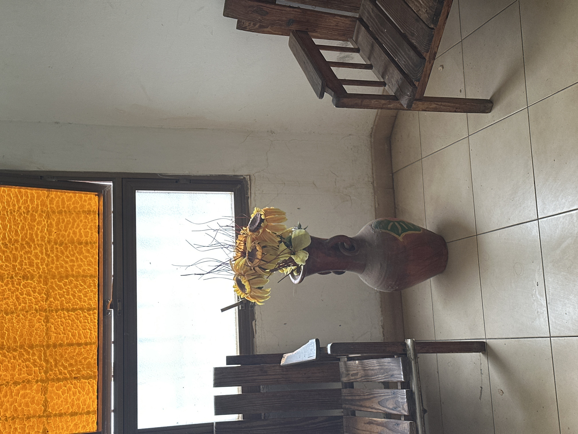 An interior scene in Juan Griego, Venezuela, depicts a corner of a room featuring a window, two wooden chairs, and a vase of artificial flowers. A window dominates the left wall, comprising an upper amber, textured glass panel, slightly ajar, and a lower rectangular pane of clear or frosted glass within a dark frame. A tall, reddish-brown earthenware vase with faint green and yellow painted motifs stands centrally before the lower window, containing artificial yellow sunflowers and bare dark branches. To the left, a rustic wooden chair with slatted back, seat, and armrest is partially visible. An identical or similar wooden chair is partially visible on the right, also featuring a slatted design and armrest. The off-white walls exhibit dirt marks, smudges, and fine cracks, particularly in the corner and near the dark tiled baseboard. The floor is tiled with light-colored square ceramic tiles and dark grout lines, showing minimal debris. Bright, diffused natural light enters through the lower window. No people or overt actions are depicted.