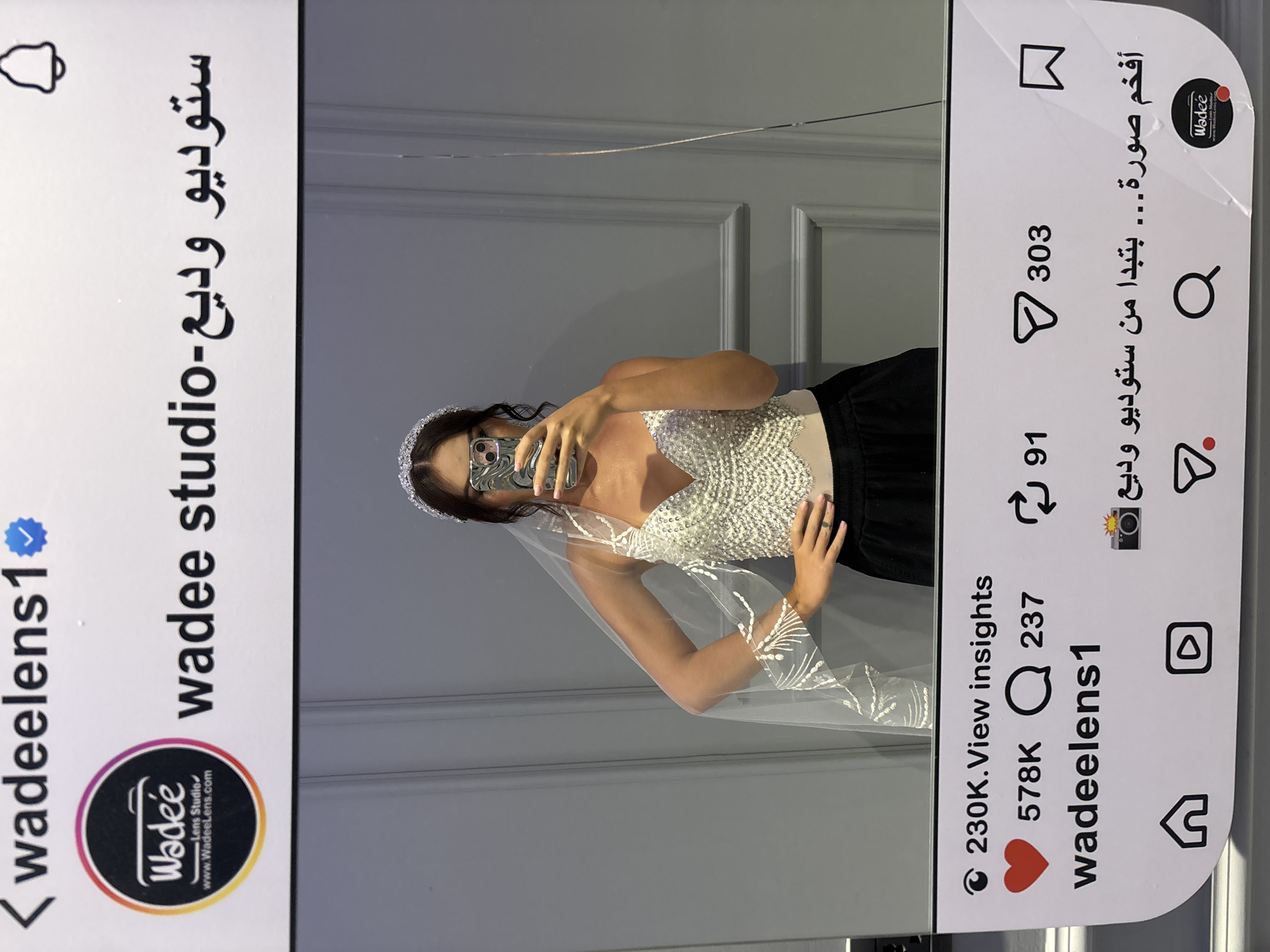 An Instagram post simulation is displayed, featuring a profile picture, username, and engagement metrics. The profile picture is a circular logo with the text "Wadee Lens Studio" and "www.Wadeelens.com". The username "wadeelens1" is visible, followed by Arabic text that translates to "Wadee studio". Below this, engagement icons show 230K views, 578K likes, 237 comments, and 303 shares. A caption in Arabic, translating to "The most amazing photo... starting from Wadee Studio", is also present, along with icons for home, play button, search, and a bookmark. The simulated post is overlaid on a photograph of a woman in a wedding veil and a sparkling top, taking a selfie in a mirror. The background is a wall with vertical paneling. This image was taken in Sweileh, Jordan.