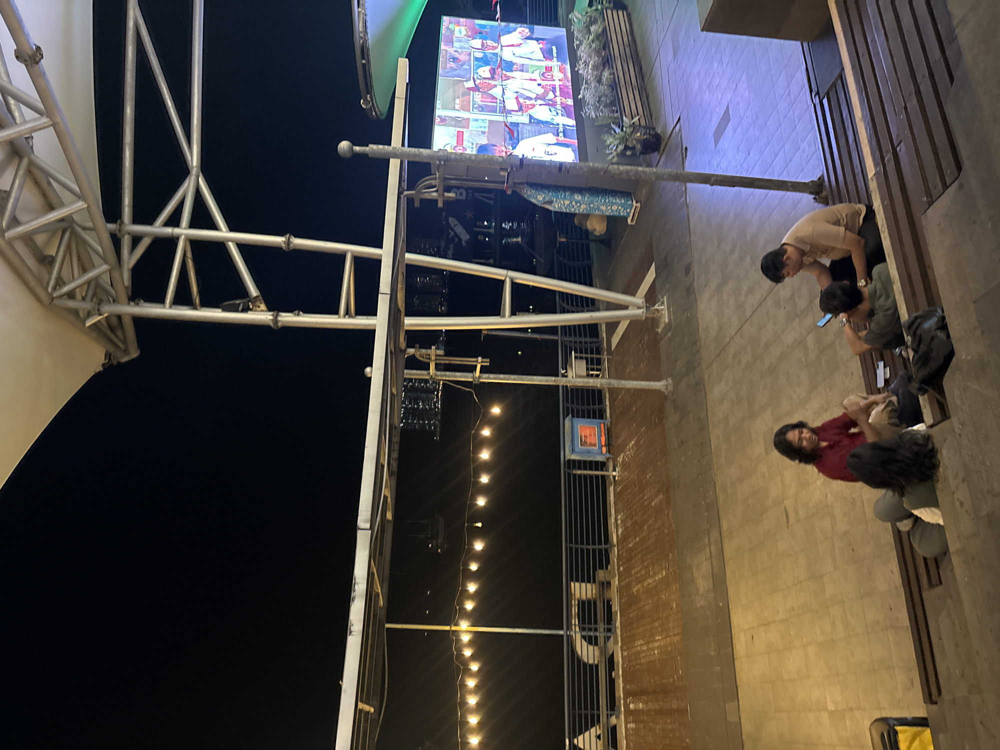 The image depicts an outdoor nighttime scene at a waterfront location in North Jakarta, Indonesia. A large screen displays images, possibly of students in uniform, situated on a platform with planters. Several people are visible, including a woman in a maroon top sitting on wooden steps, facing away from the camera, and two men also seated on the steps, one looking at his phone and the other looking down. String lights are visible along a railing overlooking a dark body of water, with city lights in the distance. A metal structure supports a canopy overhead. The ground is paved with tiles and wooden planks.
