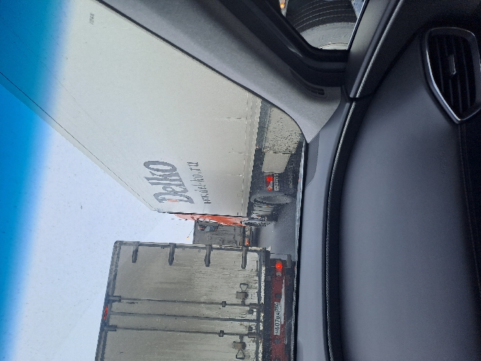 The image depicts a traffic queue of large commercial trucks on a multi-lane road in Obukhovo, Russia, observed from inside a stationary or slow-moving vehicle.
In the foreground, the dark dashboard, lower windshield, and right-side A-pillar of the observer's vehicle are visible. The dashboard features a textured surface, and a silver-accented air vent is partially seen on the far right. The top portion of the windshield shows a blue tint.
Ahead and to the left, the rear of a dirty, light-colored box truck is visible. Its two red taillights are illuminated. A Russian license plate, "H607BA|750", is affixed to its lower rear. The "750" region code corresponds to Moscow Oblast.
To the right, immediately adjacent to the observer's vehicle, is the right side and rear of a white box truck featuring "Delko" and "www.del-ko.ru" in dark blue lettering on its side. The orange cab of this truck is partially visible. Its tires and lower body are heavily soiled with dirt and grime. A partially visible Russian license plate, "H220B", is on its rear.
Further down the road, additional trucks are queued in both visible lanes. The road surface appears wet or damp. The sky is overcast, indicating a dull daytime environment. No individuals are visible within the frame.