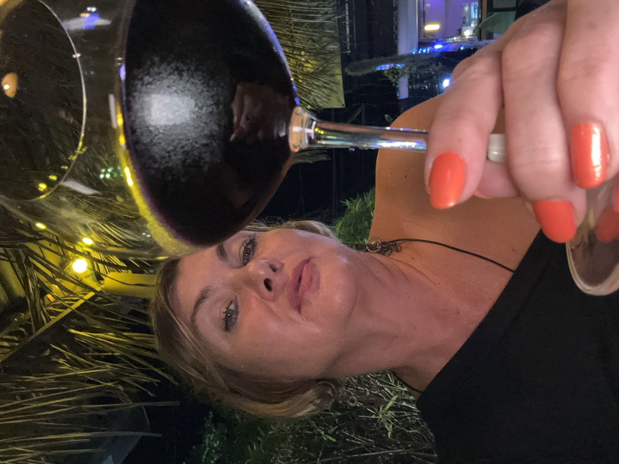 This close-up portrait, oriented vertically, captures a woman holding a wine glass at night in Baan Lamai, Thailand. The woman, with light hair and a serene expression, looks directly towards the viewer. She is wearing a dark top and a necklace with an ornate pendant. Her right hand, adorned with a ring on her ring finger, holds the slender stem of a wine glass, her nails painted a striking coral-orange color. The wine glass, filled with a dark liquid (likely red wine), is prominent in the upper left, reflecting ambient lights.
The setting is outdoors, indicated by the visible palm fronds in the background, some illuminated by warm yellow string lights. Further in the background, glimpses of blue lights and structures suggest an evening resort or outdoor dining area. The overall impression is one of a relaxed evening in a tropical location. The time of day is clearly night due to the darkness and reliance on artificial lighting. No text is visible in the image.