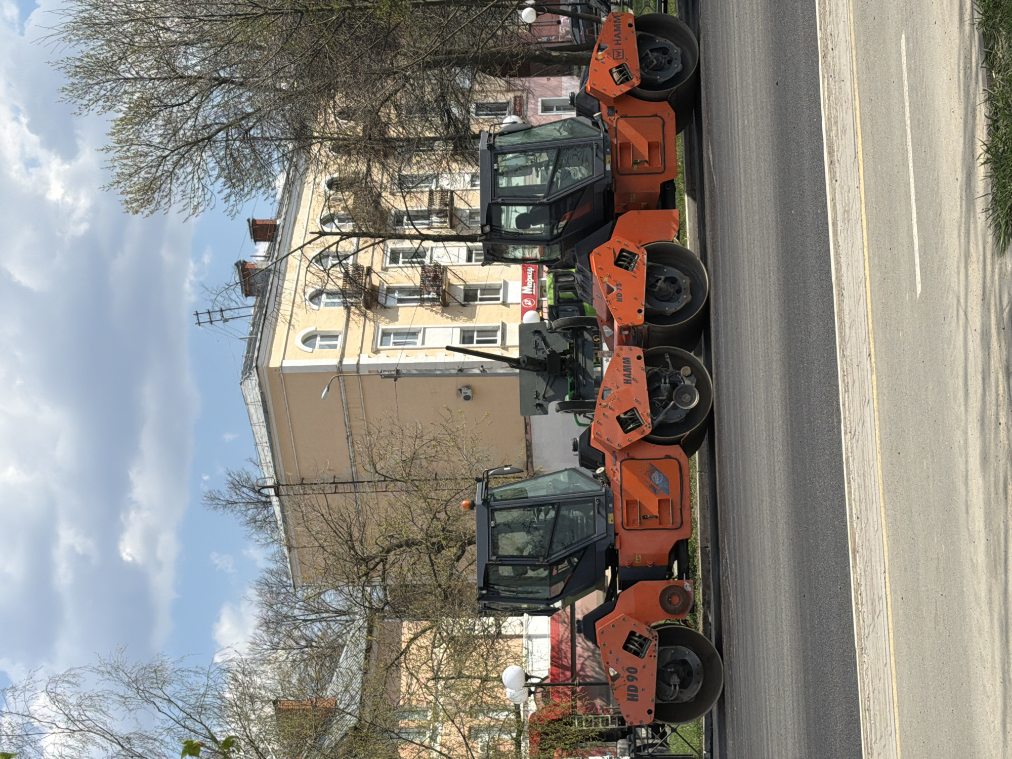The image shows a line of orange Hamm HD 90 road rollers parked along the edge of a road in Votkinsk, Russia. A multi-story yellow building with multiple windows and fire escapes stands in the background, partially obscured by bare trees. The sky is partly cloudy. The road has a painted white line dividing lanes and a yellow line marking the shoulder. Grass and some small bushes are visible on the right side of the road. There are also street lamps near the machines.