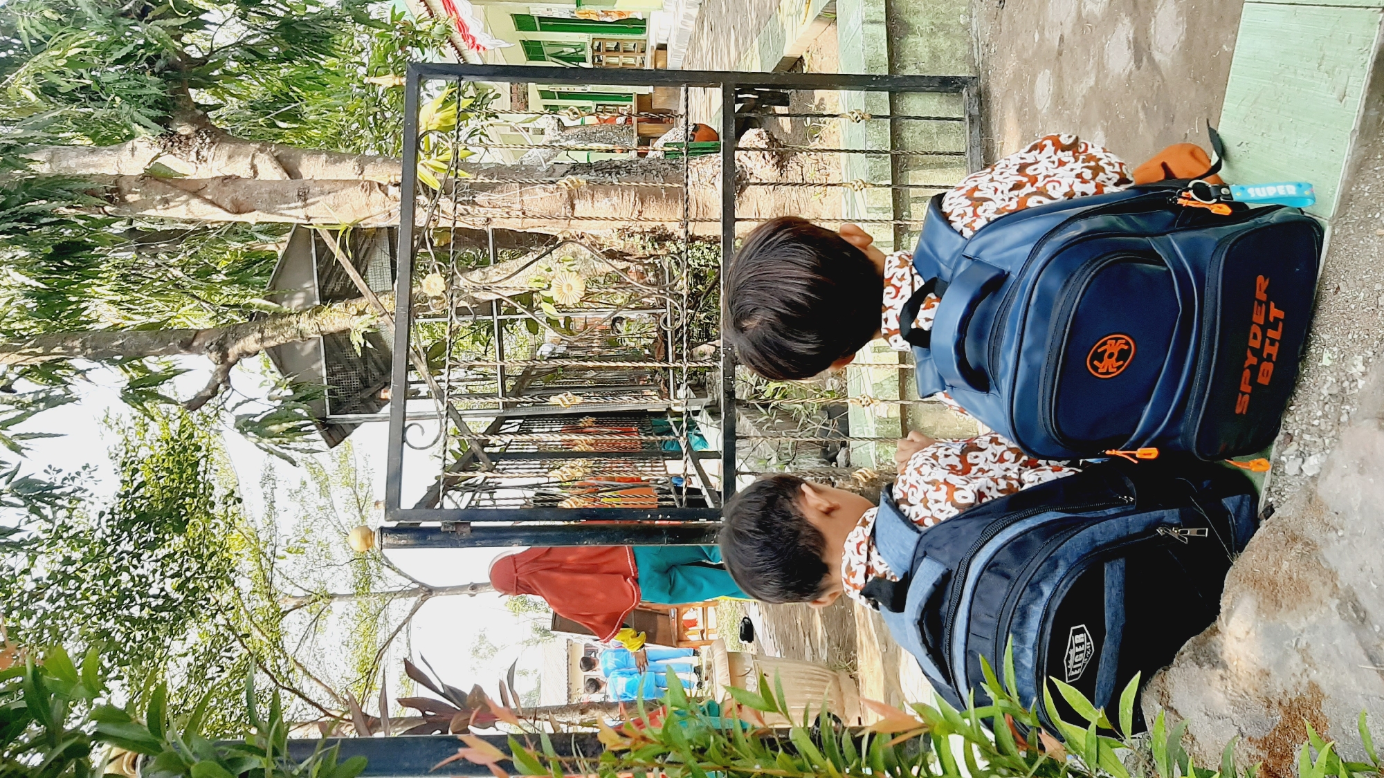 In the city of Sumberjo, Indonesia, two young boys wearing patterned shirts and blue backpacks are seated facing away from the camera, looking towards a black metal gate. Beyond the gate, a woman wearing a rust-colored hijab and teal pants stands near a whiteboard, facing three children in blue uniforms. Trees with green leaves and rough bark are prominent in the background, along with a building with green trim and white walls. The ground is covered with gravel and some stones, and a leafy plant is visible in the foreground. The scene appears to be set in an outdoor area, possibly a schoolyard, under a bright sky.