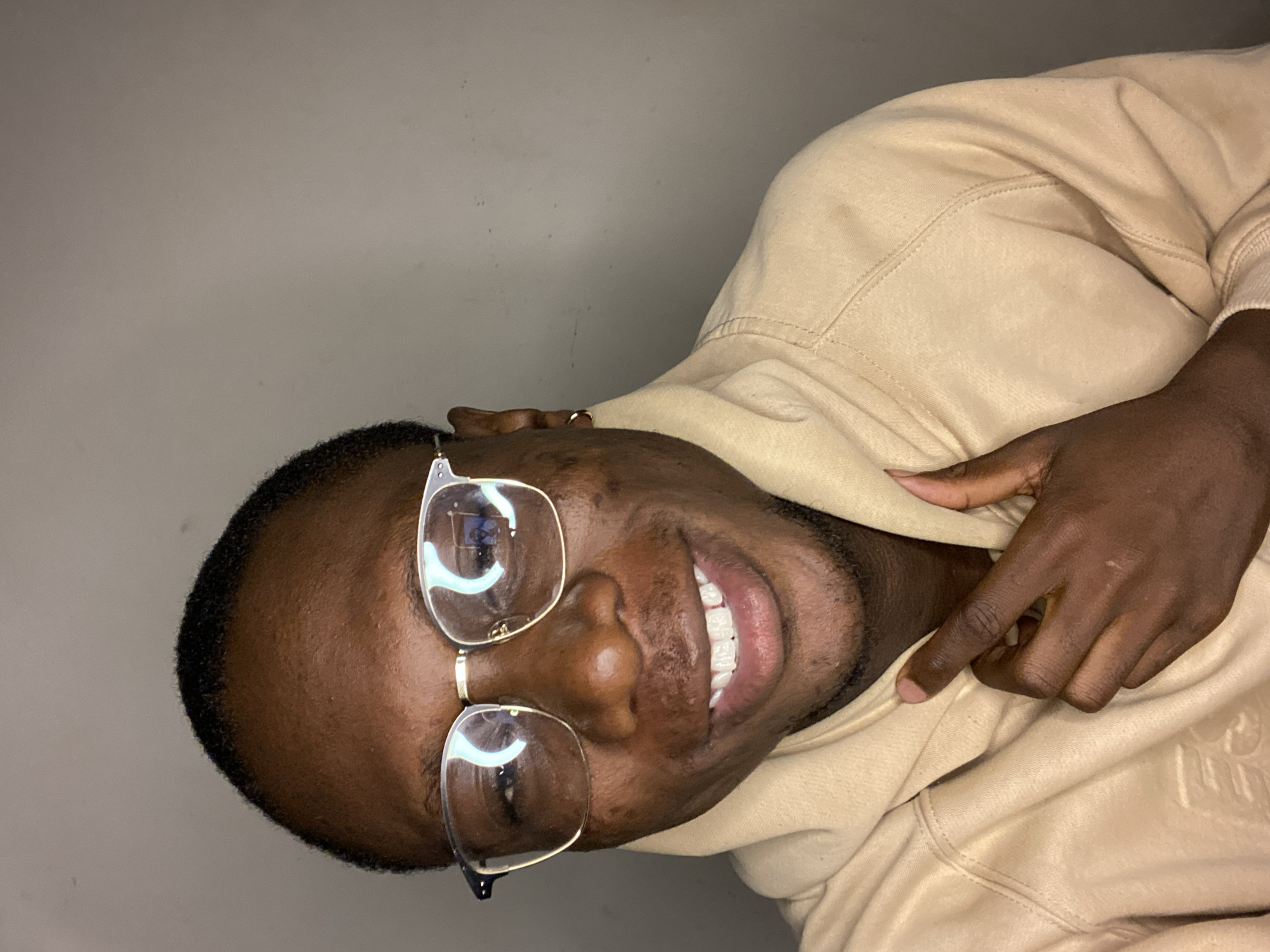 A close-up portrait features an adult individual with dark skin, short dark hair, and clear-framed glasses, smiling broadly at the camera. The person is presented as if viewing from a sideways angle, possibly due to camera orientation, but they appear upright within the frame. Their face shows a happy and friendly expression, revealing bright white teeth.
They are dressed in a light brown or tan hooded sweatshirt (hoodie), which has visible stitching details and a subtle embossed or stitched logo on the chest, though the text itself is not clearly decipherable. A gold-colored ring is worn on one of their fingers, which is gently placed on the fabric of the hoodie near their chest. The background is a plain, light gray or off-white wall, indicating an indoor setting. Reflections in the glasses suggest artificial lighting. The overall impression is one of cheerfulness and approachability. This image was captured in Bukuru, Nigeria.