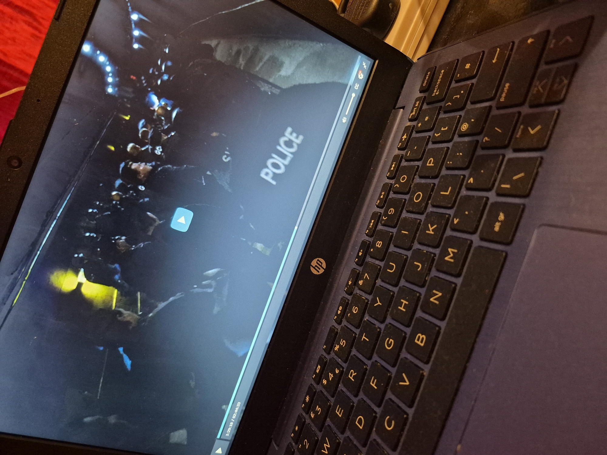A laptop is positioned in a dimly lit environment, with its screen displaying a video. The video appears to show a crowd of individuals, some of whom are wearing uniforms that resemble those of police officers, based on the visible "POLICE" text on the screen. The scene in the video is illuminated by a mix of blue and yellow lights, suggesting it might be nighttime or an indoor setting. The laptop itself is a HP brand, indicated by the logo on the keyboard. The keyboard is a standard QWERTY layout. The overall scene is captured from a slightly elevated, angled perspective, focusing on the laptop and a portion of the keyboard. There is no indication of a specific location within the city of Sandwell, United Kingdom.