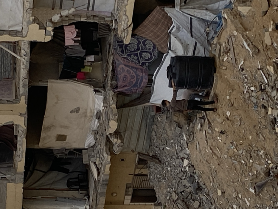 The image depicts a scene of severe structural damage in Khan Yunis, Palestinian Territory, focusing on a multi-story building and its immediate foreground. The building exhibits significant destruction, with large sections of its facade and internal walls missing, revealing exposed interiors across multiple levels. Concrete rubble and twisted rebar are visible throughout the damaged structure and on the ground.
On the upper visible floor, white tarps and fabric sheets are used to cover openings, one of which has a small rectangular patch. A black plastic chair is positioned near a draped tarp. On a lower, partially exposed floor, laundry consisting of various items, including pink, black, red, and white garments, hangs on lines. Below this, patterned blankets (purple, blue, brown) are draped to create makeshift coverings or partitions within further damaged sections.
In the foreground, a mound of sand and extensive rubble, including broken concrete blocks and debris, occupies the lower half of the frame. Two individuals are present on this mound. One individual, wearing a brown long-sleeved shirt and dark pants, stands facing right, with their right hand resting on top of a large, black, cylindrical plastic water tank. A second individual is partially visible behind the first, obscured by the water tank, appearing to be in a stooped or kneeling posture near the base of the tank. The environment is dominated by remnants of destroyed buildings and scattered debris.
