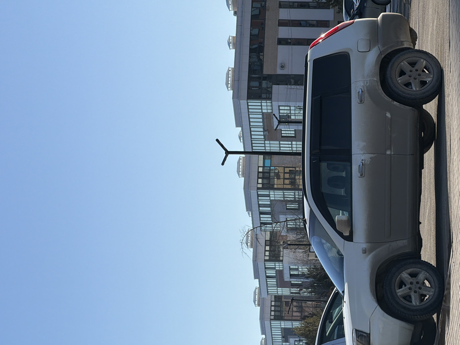 The scene depicts a daytime outdoor urban environment in Dudareva, Russia, under a clear, bright blue sky. In the midground, a light-colored (champagne or beige) Nissan X-Trail SUV, likely a first-generation model, is parked facing left. The vehicle exhibits significant accumulation of dirt or dust along its lower body panels, fenders, and wheels. Its passenger-side windows and rear window are dark-tinted. Sunlight is prominently reflected off the vehicle's rear right taillight. A partial view of another light-colored vehicle is visible in the foreground to the left. In the background, a uniform row of modern, multi-story buildings with light-toned facades and large, reflective bluish-green windows extends across the horizon. These buildings feature flat roofs with small structural elements and visible exterior air conditioning units. Several black, Y-shaped streetlights are positioned across the scene, one prominently in the midground. Sparse, bare-branched vegetation is visible between the buildings. No individuals are present within the frame, and no dynamic actions or interactions are occurring.