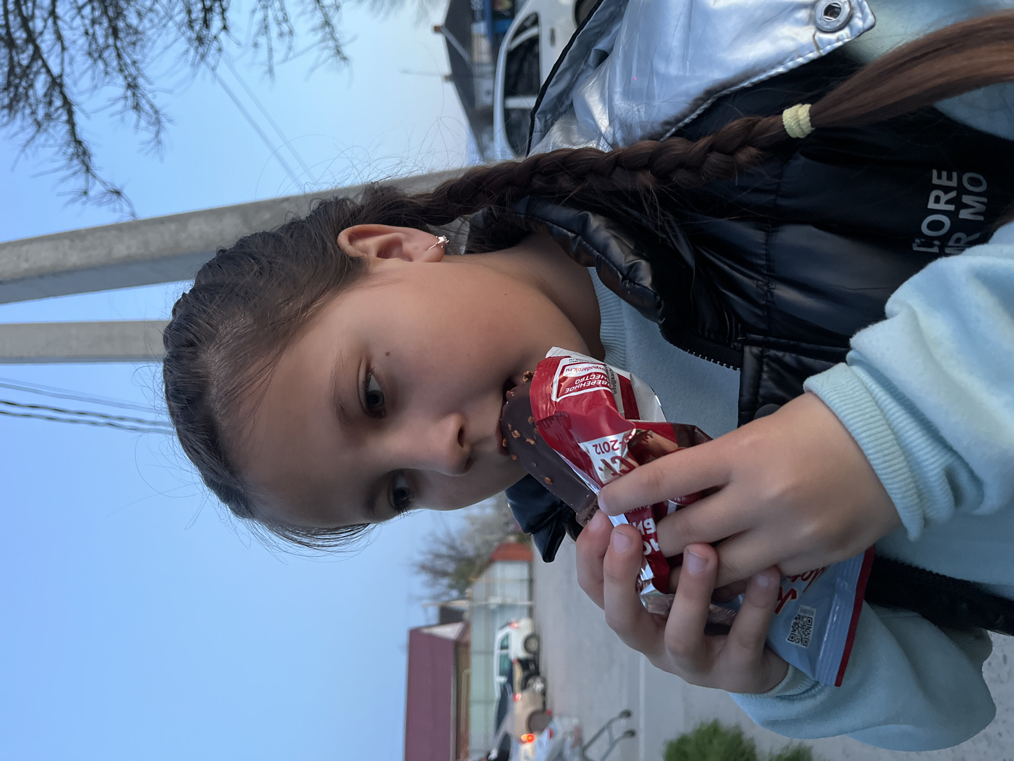 A young girl is captured in a close-up shot, eating a chocolate-covered ice cream bar with nuts. She has long, dark hair styled in a braid secured with a yellow elastic band, and a small star-shaped hoop earring is visible on her right ear. She wears a shiny silver and black puffer jacket over a light blue sweatshirt. Her attention is focused on the ice cream she holds in both hands, with a bite taken from the top.
The setting appears to be outdoors in Novopokrovskaya, Russia, possibly on a street or roadside. The sky is a clear, light blue, suggesting either late afternoon or early evening. In the background, there's a concrete utility pole, bare tree branches, and overhead power lines. Further back, distant buildings, including a house with a red roof, and a white vehicle are visible.
Visible text on the ice cream wrapper includes Cyrillic words such as "БЕРЕНДЕЕВО" (Berendeyevo), "ВКУС" (Taste), and "МЫС" (Cape/Thought), along with the number "2012" and a QR code. Part of a brand name, "CORE" and "MO," is visible on the black lining of her jacket. The overall atmosphere is casual and relaxed, with the girl enjoying her treat.