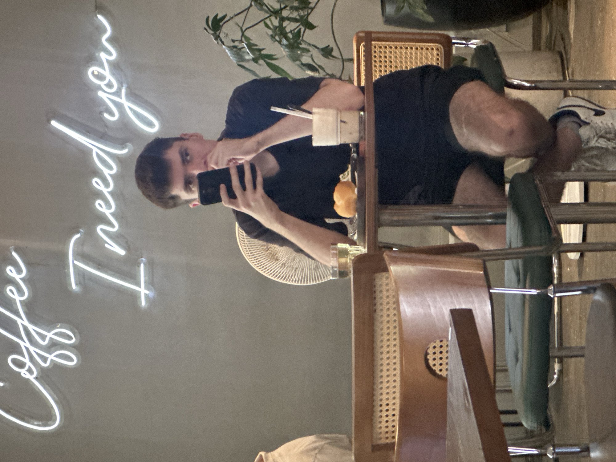 A man is sitting at a table and taking a photo with his phone. He is wearing a black t-shirt and black shorts. The table has a drink on it. In the background is a neon sign that says "Coffee I need you". The location is likely a café or coffee shop. The city of Bac Nha Trang Ward, Vietnam, is not apparent from the image.