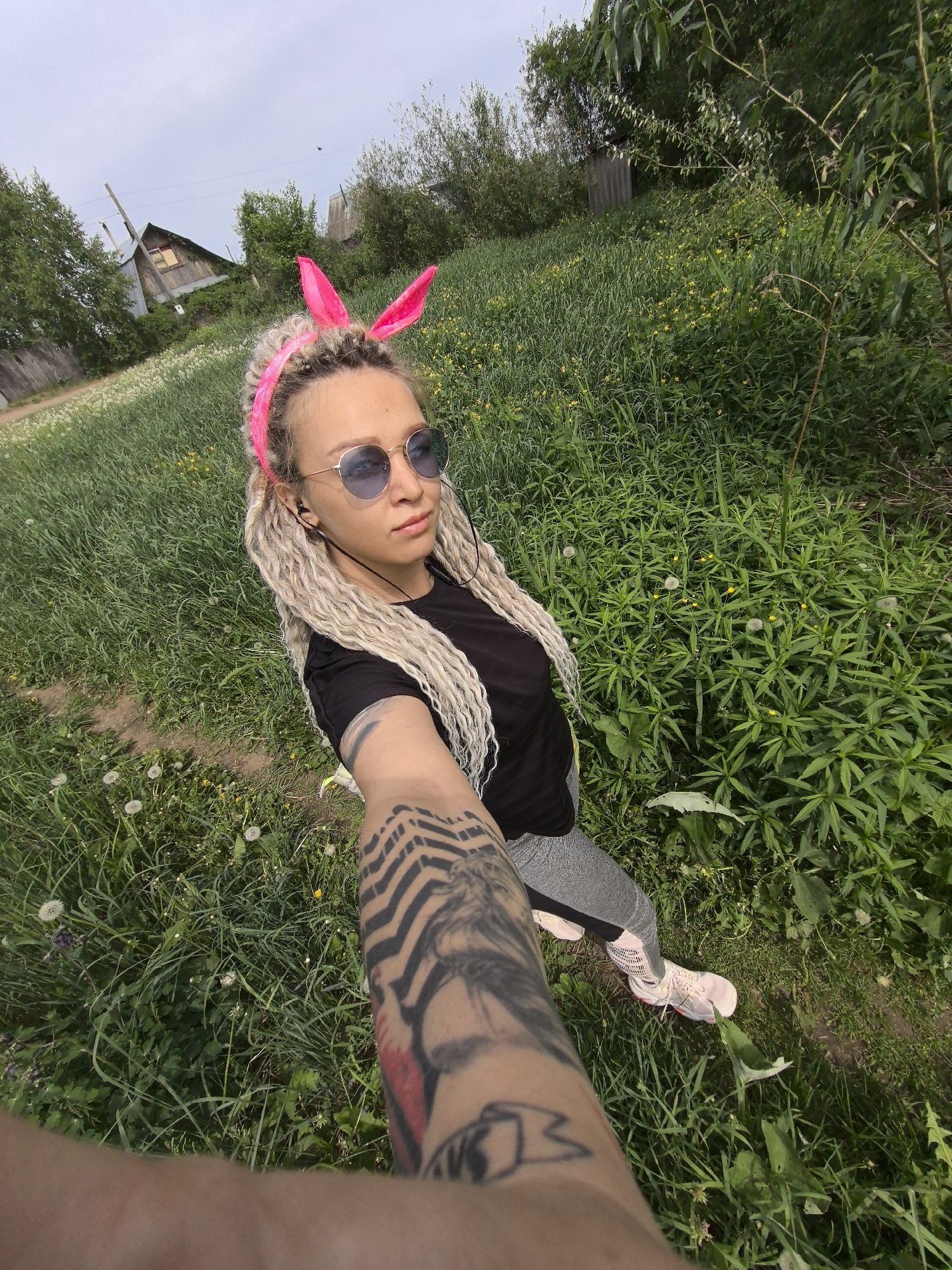 A woman takes a selfie outdoors. She is wearing a black t-shirt, grey pants, pink sneakers, and sunglasses. She has blonde dreadlocks and pink bunny ears. Her arm is extended towards the camera, displaying tattoos. The background includes green grass, plants, and a wooden house in the distance. The photograph was taken in or near Noviy, Russia.