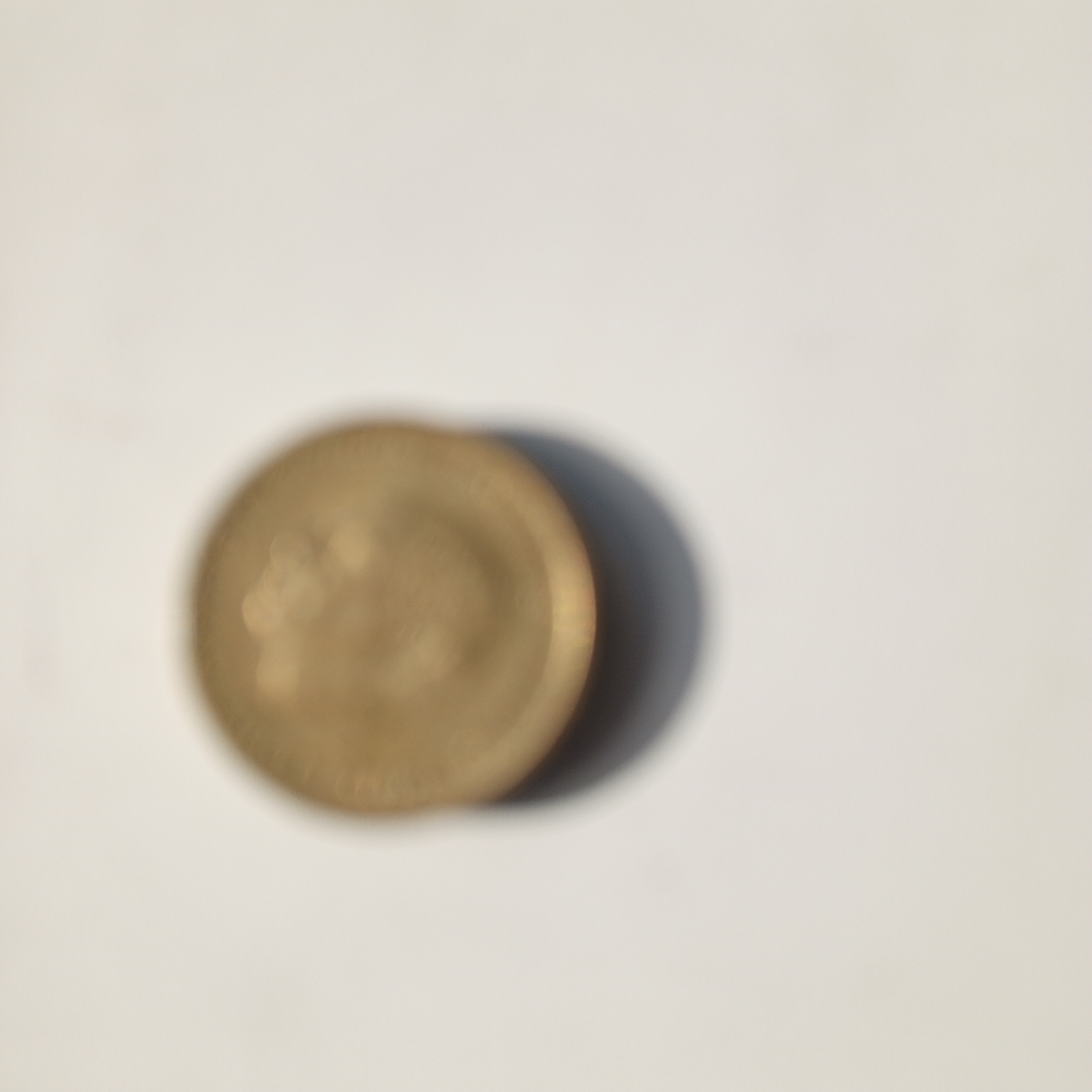 A single, out-of-focus, gold-colored coin is visible on a white, flat surface. The coin casts a shadow to its right. The image does not contain any information that would allow for its location to be identified as Sayanogorsk, Russia.