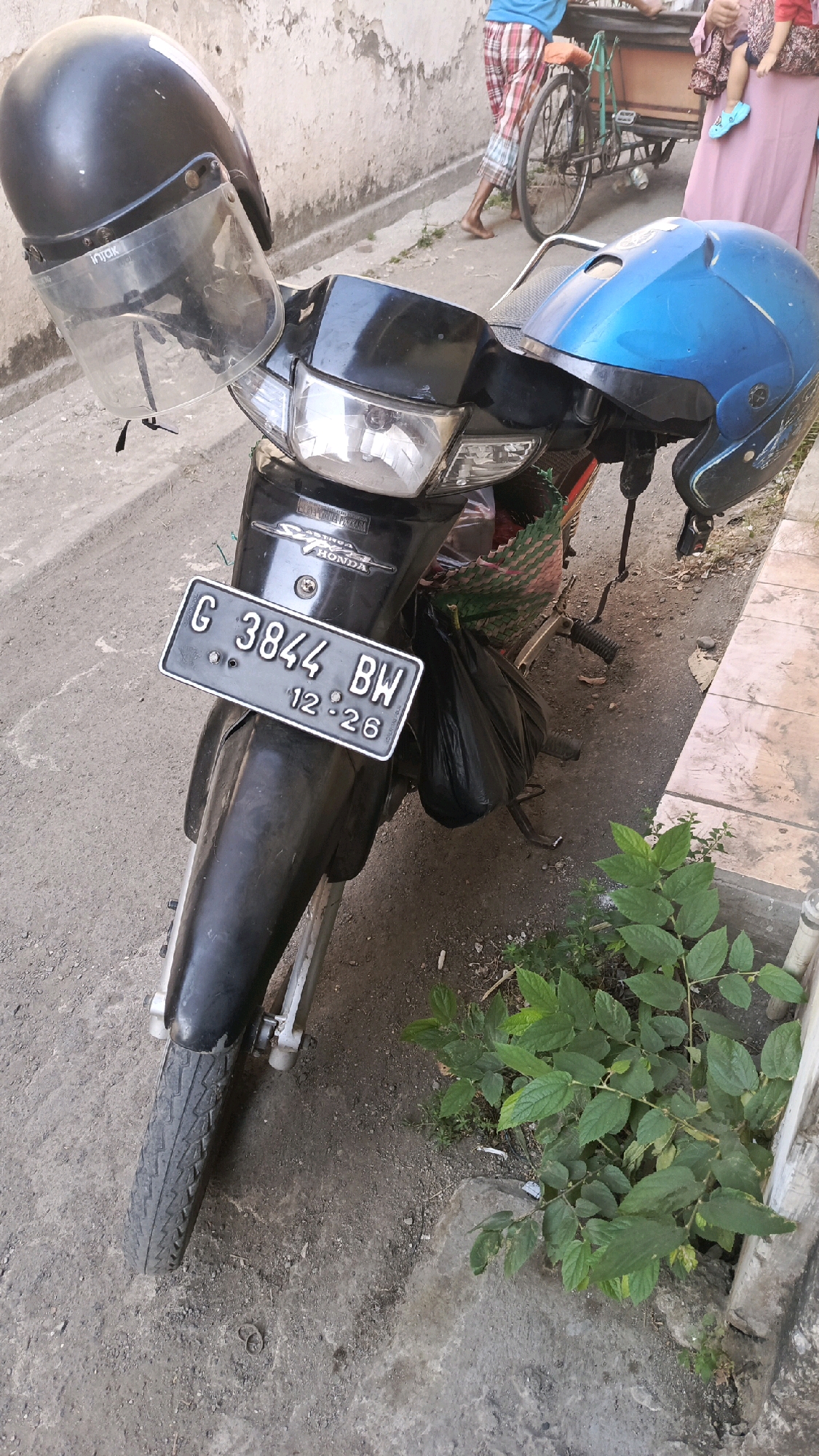 In Pekauman Kulon, Indonesia, a black motorcycle is parked on a dirt and concrete surface, with a black helmet resting on its handlebars and a blue helmet on the seat. The motorcycle's license plate reads "G 3844 BW 12.26". In the background, a person wearing plaid shorts and a blue shirt is pushing a bicycle with a cart. Another person, partially visible, is holding a baby wearing a pink outfit. Green plants grow in the foreground, and a building with a textured wall is visible in the background.