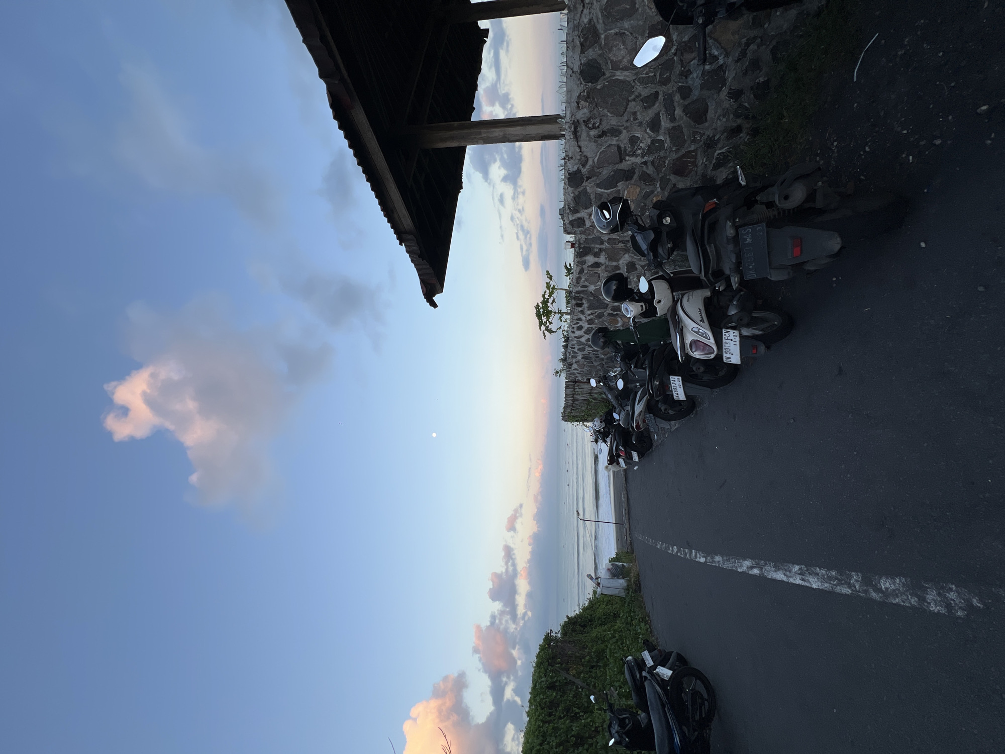 The image depicts a row of parked motorbikes along a road in an area near the ocean. The sky is visible above, exhibiting a gradient of colors indicative of dawn or dusk, with clouds scattered throughout. Adjacent to the road, there's a stone wall and some vegetation. The location is suggested to be in the city of Unknown, Indonesia.