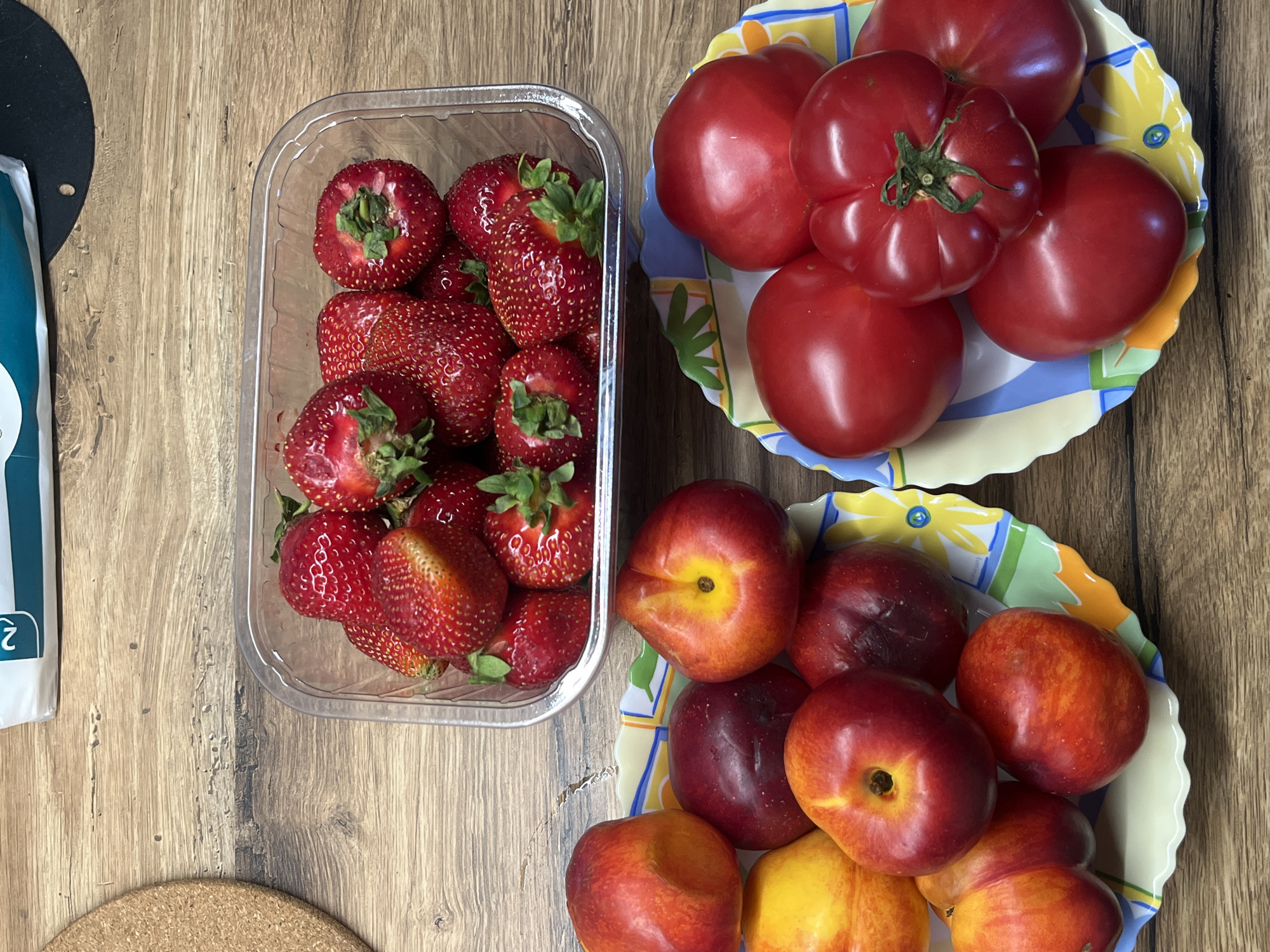 On a wooden surface, two decorative plates are visible, one above the other. The top plate holds several nectarines and what appear to be plums, with colors ranging from yellow to deep red. The bottom plate contains several bright red tomatoes, one of which has a prominent green stem. To the right of the plates, a clear plastic container is filled with fresh strawberries. The background is a simple wooden table. To the far right, a portion of a white and teal package is visible, with the number "2" printed on it. The scene depicts a collection of fresh produce arranged on a table.