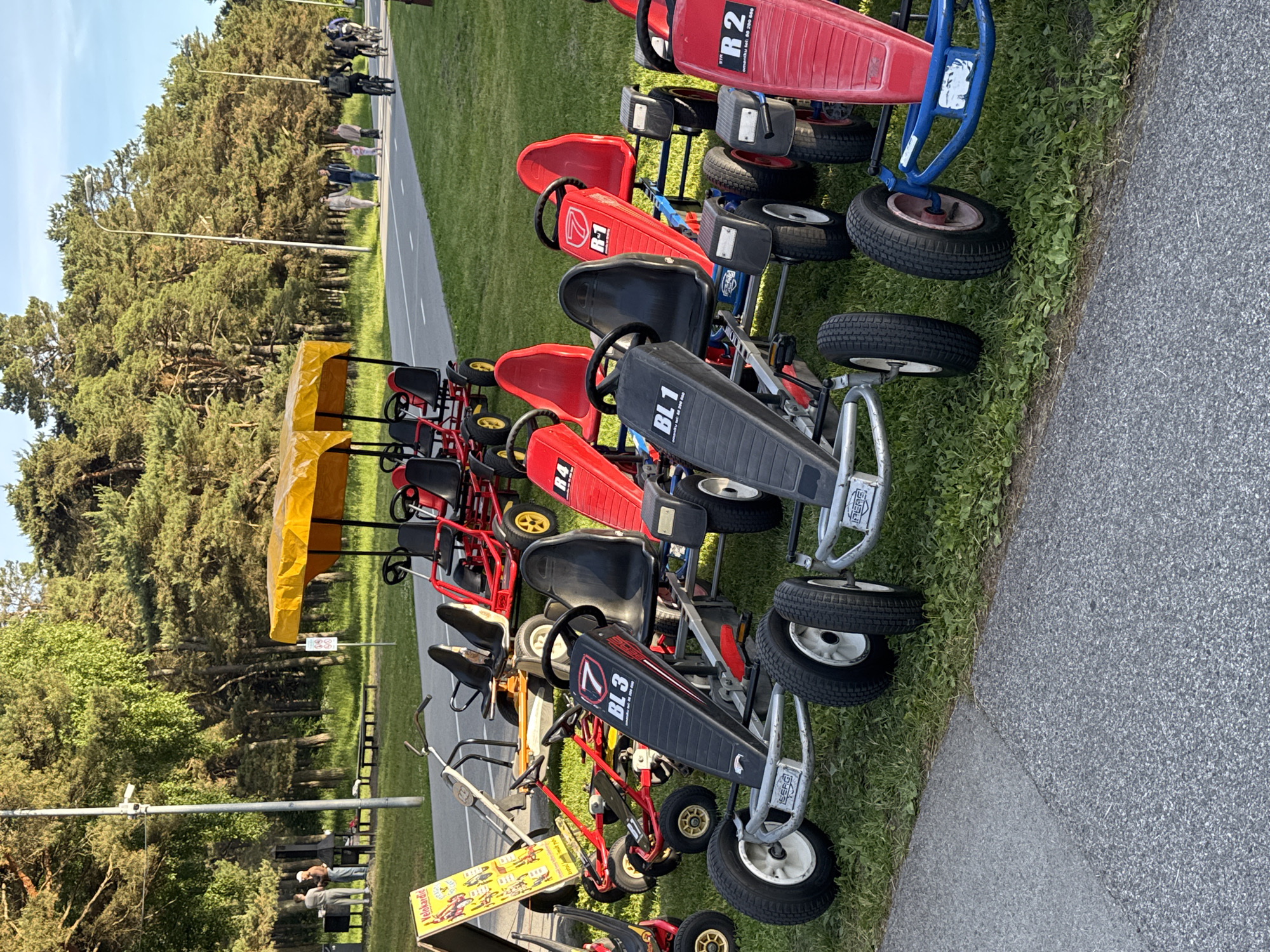 In Tallinn, Estonia, a row of pedal go-karts are parked on a grassy area next to a paved path. The go-karts are of various colors, including red, black, and yellow. Some of them are double-seated, while others are single-seated. A yellow canopy covers some of the go-karts. In the background, there are trees and a few people walking and cycling on the path. The scene suggests a recreational area, possibly a park or a promenade by the sea. The overall atmosphere is relaxed and summery.