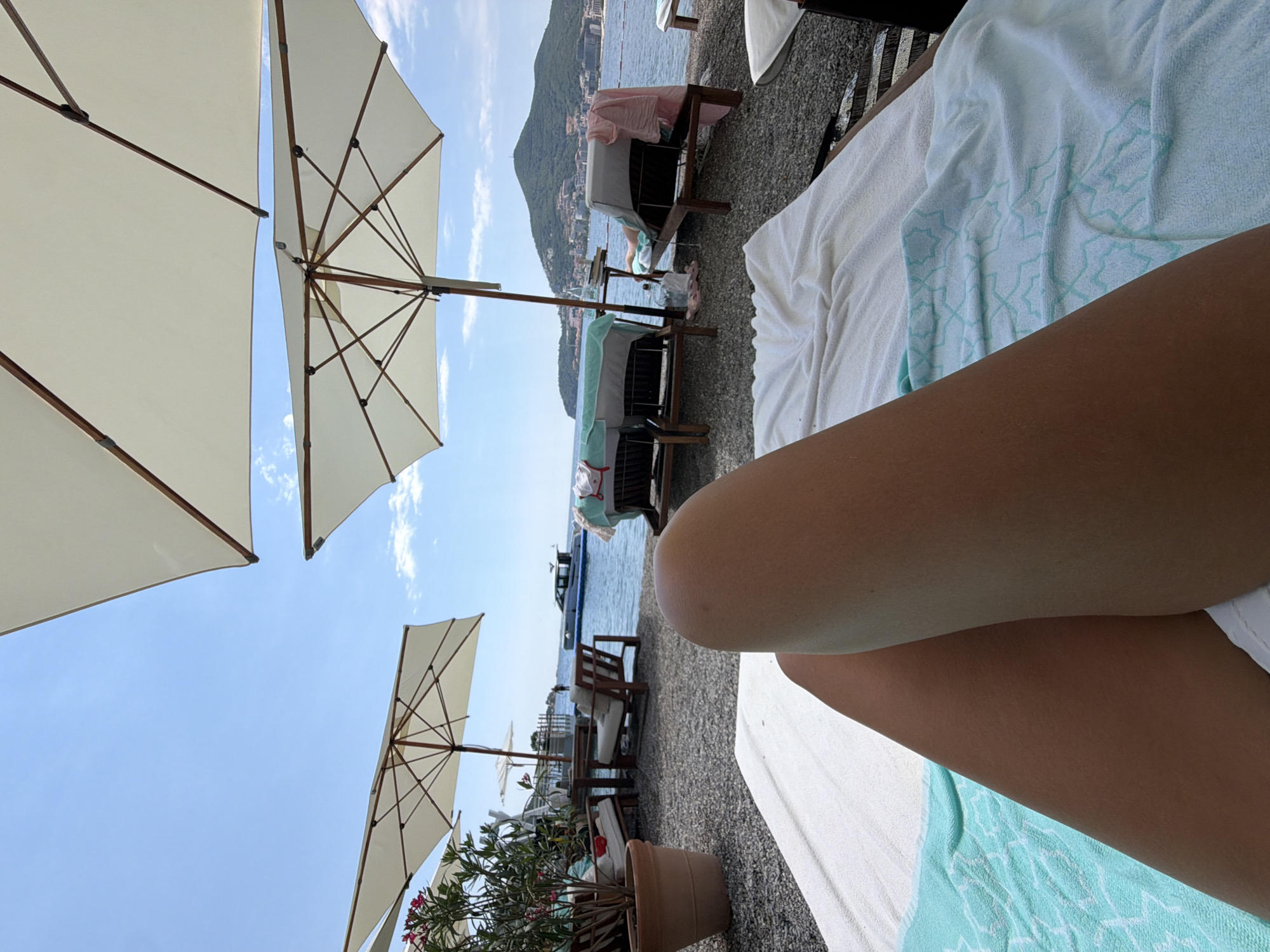 This is a first-person perspective shot of a person lying on a sun lounger at a beach resort. The subject's legs are prominently in the foreground, resting on a white towel with a light blue geometric pattern. In the background, there are several white beach umbrellas, sun loungers with towels, and lounge chairs. The beach is covered in pebbles. The sea is visible, with a white boat cruising in the distance. A lush green hill covered with buildings forms the backdrop. The sky is clear blue with a few scattered clouds. This scene appears to be from the Budva Riviera in Montenegro.