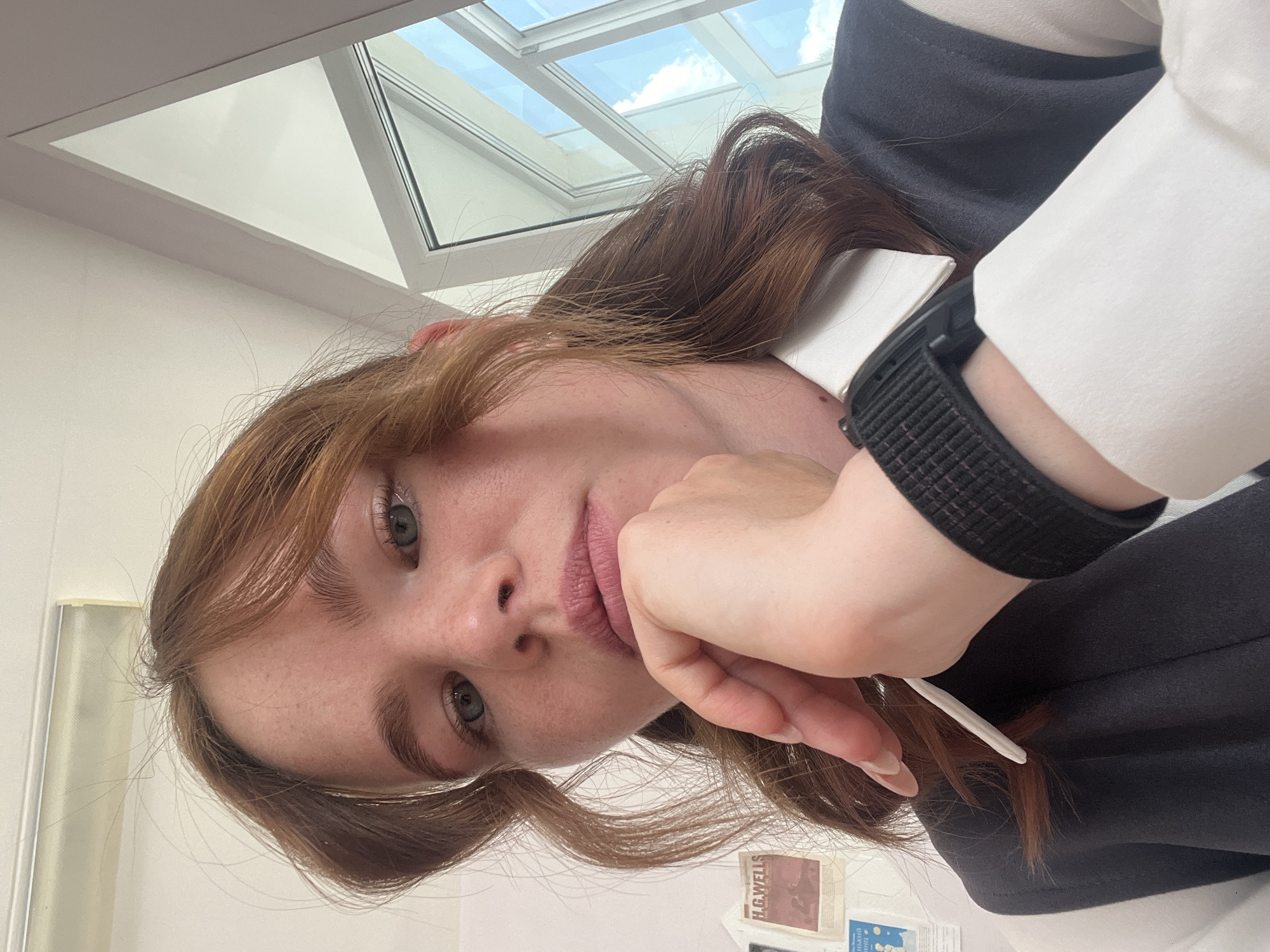 A young woman with auburn hair and light blue eyes is captured in a close-up, overhead shot. She is resting her chin on her hand, with her arm propped up. She is wearing a white collared shirt and a dark grey vest or jacket. A black smartwatch is visible on her left wrist. Her hair is long and falls loosely around her face and shoulders.
The background features a skylight with a view of a bright blue sky with scattered white clouds, suggesting it is daytime and the weather is clear. The interior walls are a light, neutral color. A small part of a shelf with some items is visible at the bottom right, including a book with the text "H.G.WELLS" clearly legible.
The woman's expression is neutral, with her lips slightly parted. The overall mood of the image appears relaxed and contemplative. The image is oriented sideways, suggesting it was taken with a phone. The lighting indicates it's daylight. No specific activity or event is explicitly depicted, other than the woman posing for a photo. The location context of Chaikovsky, Russia is provided, but there are no specific visual cues within the image to confirm this location.