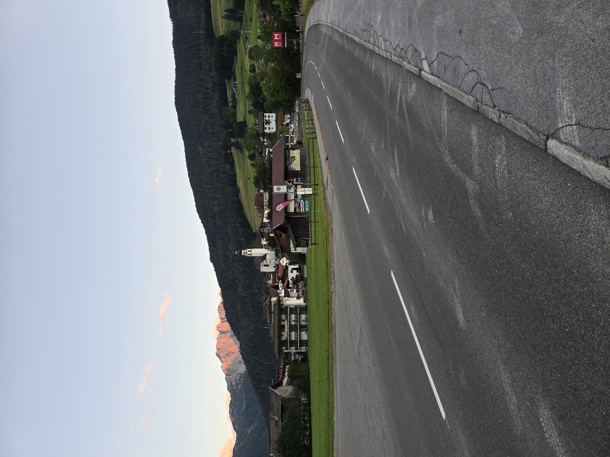 The image shows a view of Lermoos, Austria, with the Zugspitze mountain range illuminated by the setting sun in the background. In the foreground, there is a paved road with a white line marking the lanes. To the right of the road, there is a sidewalk with a cracked curb. Beyond the road, there is a green field with some trees and buildings. The buildings appear to be part of a village, with a church spire visible in the distance. The sky is clear with a few light clouds. The overall scene depicts a peaceful evening in the Austrian Alps.