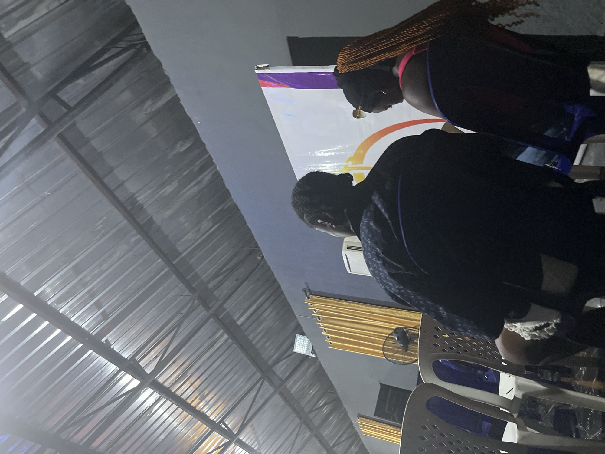 The image shows the interior of a venue with a corrugated metal ceiling. In the foreground, two individuals are seated on plastic chairs, viewed from behind. The person on the left wears a dark patterned shirt, and the person on the right wears a pink top and has long, braided blonde hair. They are facing a backdrop with a large graphic design featuring orange, purple, and white elements. To the left of the backdrop, a yellow curtain is visible, and behind the chairs, a standing fan is positioned. The environment appears to be dimly lit, suggesting an indoor event or gathering.
