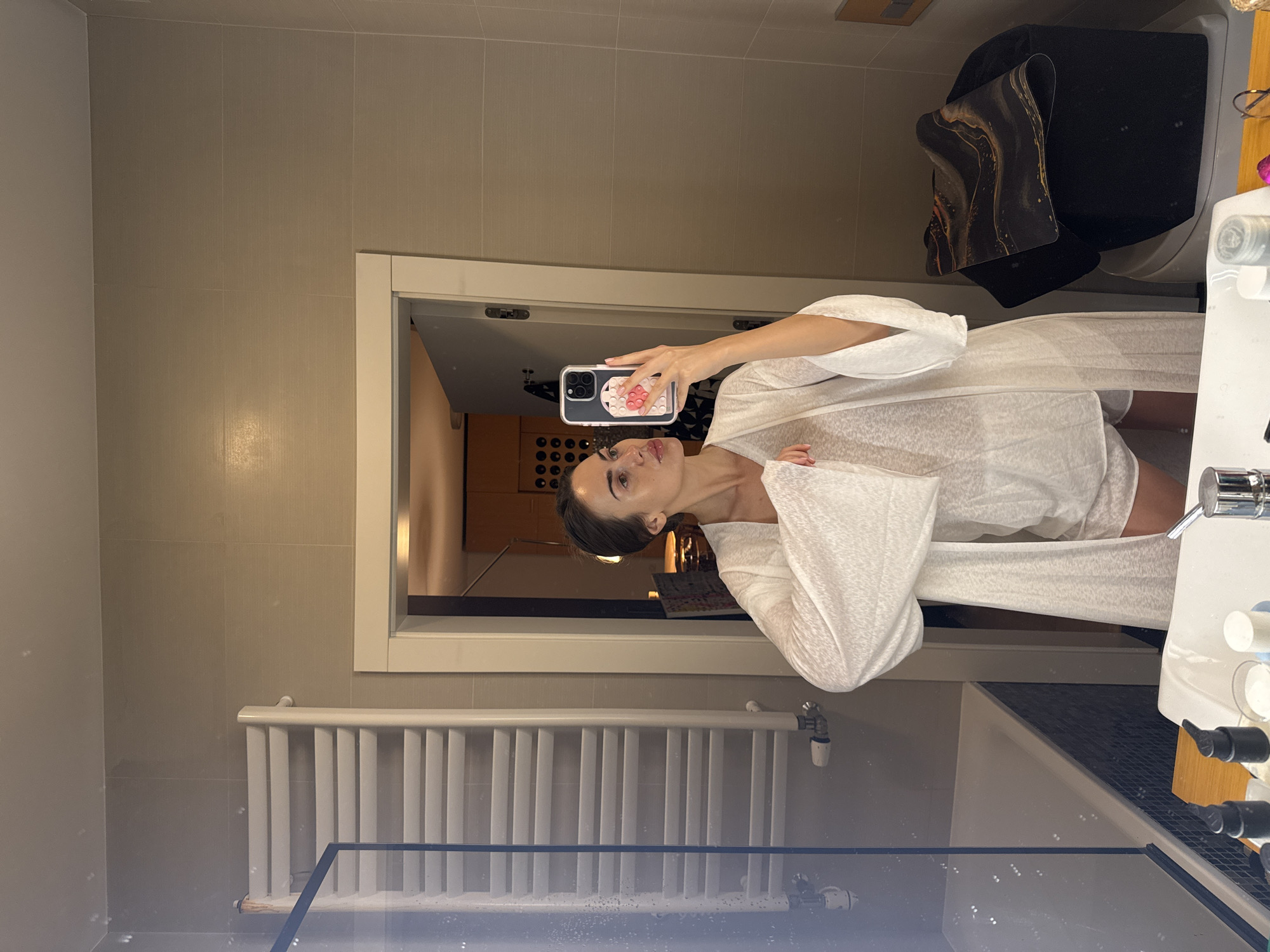 A woman is taking a selfie in a bathroom. The bathroom has tiled walls, a radiator, a shower cubicle, and a vanity with a sink and toiletries. The reflection in the mirror shows a hallway leading to what appears to be a kitchen. The location appears to be in Warsaw, Poland.