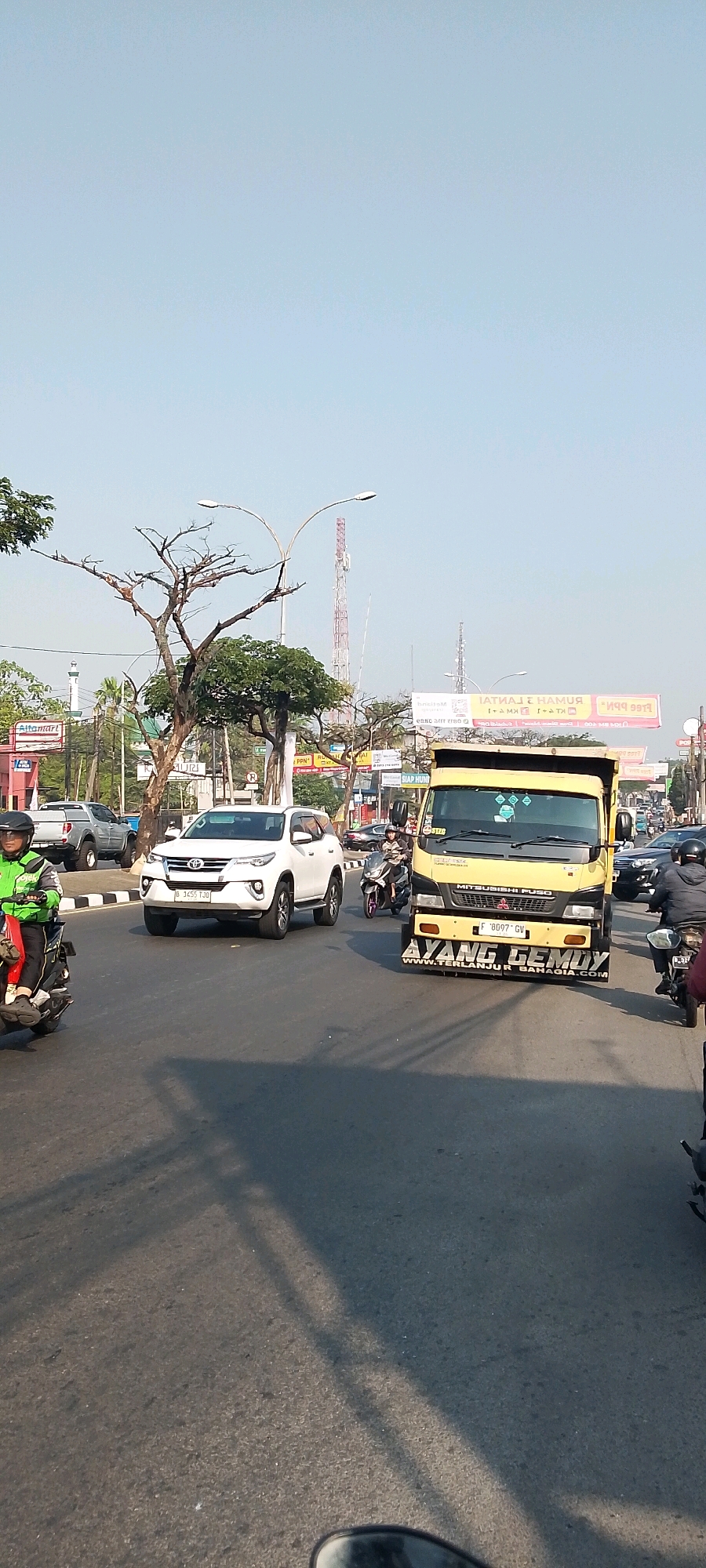 The image captures a daytime scene on a road in Gunung Putri, Indonesia, likely a busy commercial or industrial area given the presence of numerous vehicles and advertisements. The sky is clear and pale blue. In the foreground, a dark asphalt road stretches into the distance, with shadows cast across it, suggesting the sun is positioned to the left.
Several vehicles are present. A white Toyota Fortuner SUV is prominently positioned in the left-center of the frame, facing right. To its left, a motorcycle with a rider wearing a green jacket and black helmet is also moving right. Further to the right, a bright yellow Mitsubishi Fuso dump truck is driving towards the viewer, with the words "AYANG GEMOY" and "WWW.TERLANJURBAHAGIA.COM" visible on its lower front panel. A motorcycle with two riders is visible behind the yellow truck. Several other vehicles, including cars and motorcycles, are present in the background, creating a sense of traffic.
The environment includes roadside elements typical of a commercial area. On the left, behind the SUV, a tall white tower structure, possibly a minaret or part of a building, is visible. Further left, a red and white sign for "Alfamart" can be seen, indicating a retail presence. Several trees, some bare and skeletal, and others with green foliage, are scattered throughout the scene. In the background, utility poles with streetlights and telecommunication towers are visible. Billboard advertisements are also present along the roadside, one featuring reversed text that reads "HAMUR LATIHAN" and another with "MUMPU" and "free ppn". The overall impression is one of an active and bustling urban thoroughfare.