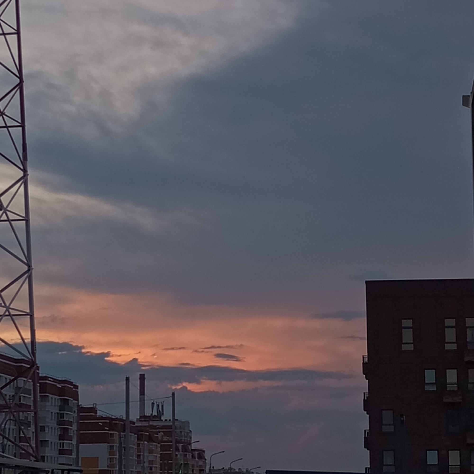 A cityscape is depicted during twilight, with a large, dark building dominating the right side of the frame and a metallic tower structure on the far left. The sky above displays a gradient of deep blue and grey clouds, with a band of warm orange and pink hues near the horizon, indicating a sunset. Several multi-story residential buildings, characterized by their brick and white facades, are visible in the mid-ground. The scene appears to be located in Pervomaysky, Russia, as suggested by the presence of apartment blocks typical of Russian urban environments and the characteristic industrial tower. No people are visible in the image, and no specific actions or interactions are taking place. The overall atmosphere is quiet and serene, with the twilight colors creating a dramatic backdrop for the urban structures.