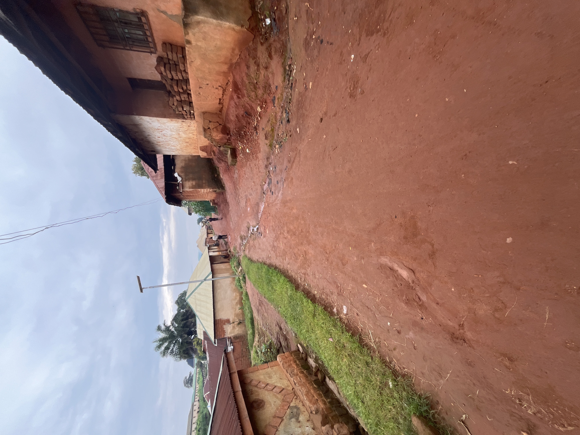 This image depicts a rural village road in Gembu, Nigeria, under an overcast sky. The road is unpaved and composed of reddish-brown soil, appearing wet and somewhat muddy in places, with scattered debris. On either side of the road are traditional houses with walls made of mud or brick and roofs of corrugated metal. Some houses are single-story, while others appear to be part of a larger compound. There is a narrow, overgrown drainage ditch running along the left side of the road. In the distance, two figures are walking away from the camera, and a street lamp is visible. A palm tree and other vegetation are also present. The overall scene suggests a quiet, ordinary day in a rural Nigerian community.