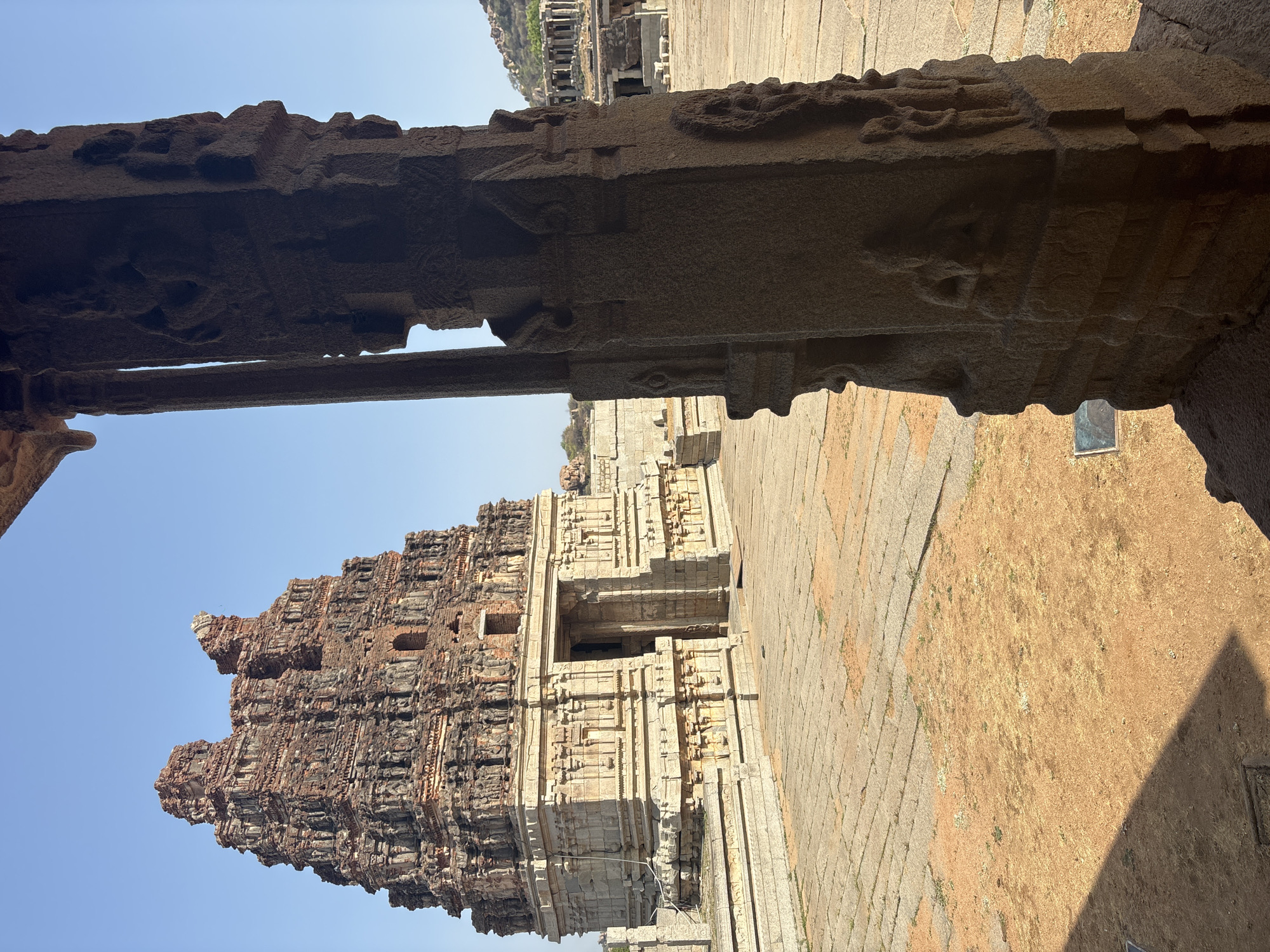 The image shows a portion of the Virupaksha Temple in Hampi, India, as seen from inside the temple complex through an ornately carved stone structure. The temple's eastern gopuram (tower) dominates the view, rising against a clear blue sky. The gopuram is a towering pyramid-shaped structure with multiple layers of intricately carved tiers, predominantly brick-red and pale stone. The structure features a succession of diminishing stacked stories, replete with carvings of deities and architectural details. The foreground includes a close-up view of a stone beam and pillar adorned with detailed carvings, partially framing the view of the tower. The floor and ground are made of large stone slabs, typical of the architecture in Hampi. Adjacent to the stone floor, there is a patch of dry, yellow grass and dirt. The scene is bathed in bright sunlight.