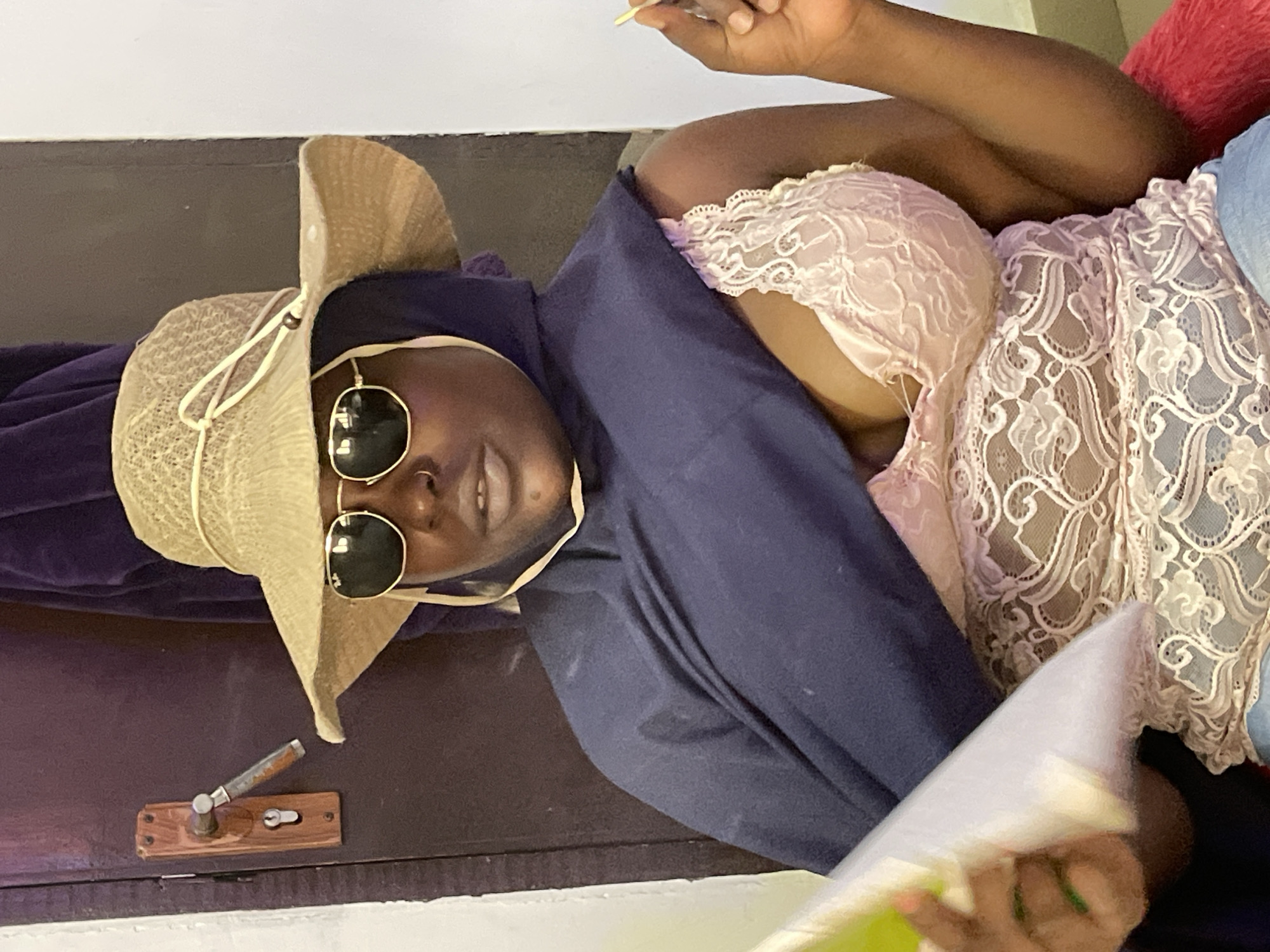 A dark-skinned woman is reclining indoors in Gwarinpa, Nigeria. She is wearing a floppy straw hat and sunglasses, a purple hooded robe, a light pink lace bra, and a light blue lace top. She is holding a piece of paper in her hands. A wooden door with a metal handle is visible in the background.