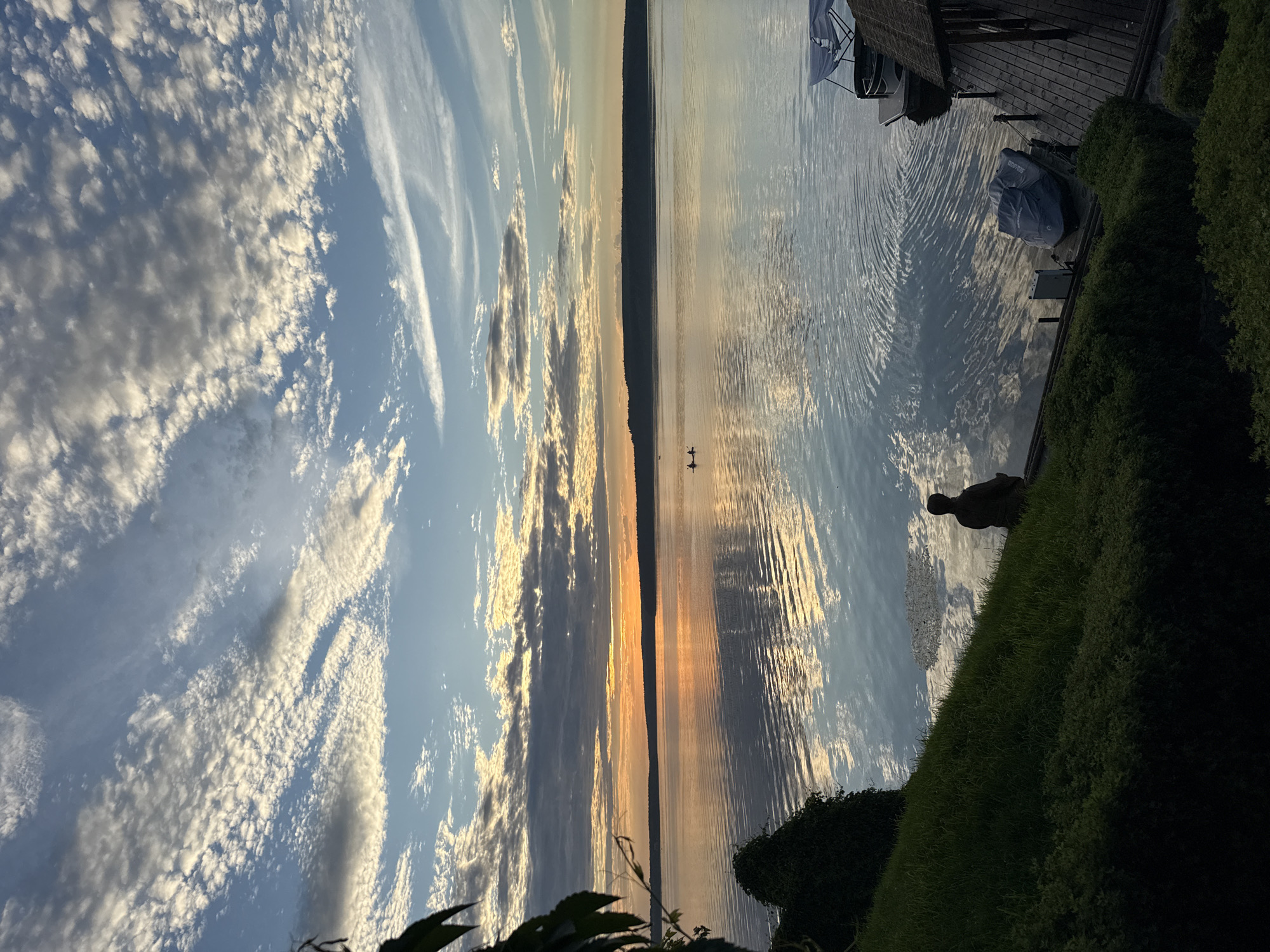 The image depicts a serene sunset over a tranquil body of water, likely a lake or large river, with a distant treeline on the horizon. The sky is a canvas of blues and oranges, adorned with scattered, fluffy white clouds that reflect beautifully on the water's surface. In the foreground, a person is seated on the grassy bank, facing the sunset. To the right, a wooden dock extends into the water, with a covered boat and a jet ski docked beside it. The overall atmosphere is peaceful and quiet, suggesting a calm evening by the water.
The location of Votkinsk, Russia, is not visually identifiable from the image itself.