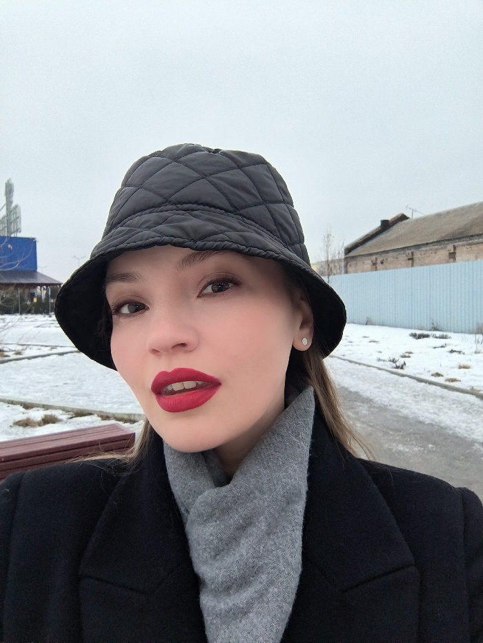 A female subject is depicted in a close-up, chest-up shot, captured outdoors in Vinnytsia, Ukraine. The subject is a young adult female with fair skin, dark eyebrows, and brown eyes, looking directly towards the camera. She wears a black quilted bucket hat, a black coat, and a grey knitted scarf. Her lips are coated with bright red lipstick, and a small, circular, light-colored stud earring is visible in her right earlobe. The background shows a winter environment with patches of snow covering the ground. A dark red wooden bench is partially visible in the lower-left. A paved pathway runs along the right side of the frame. In the mid-ground, on the left, a building with a blue facade and a multi-mast structure is visible. On the right, a light blue corrugated metal fence stands in front of an older, light-brown brick building with a dark roof. The sky is overcast and grey.