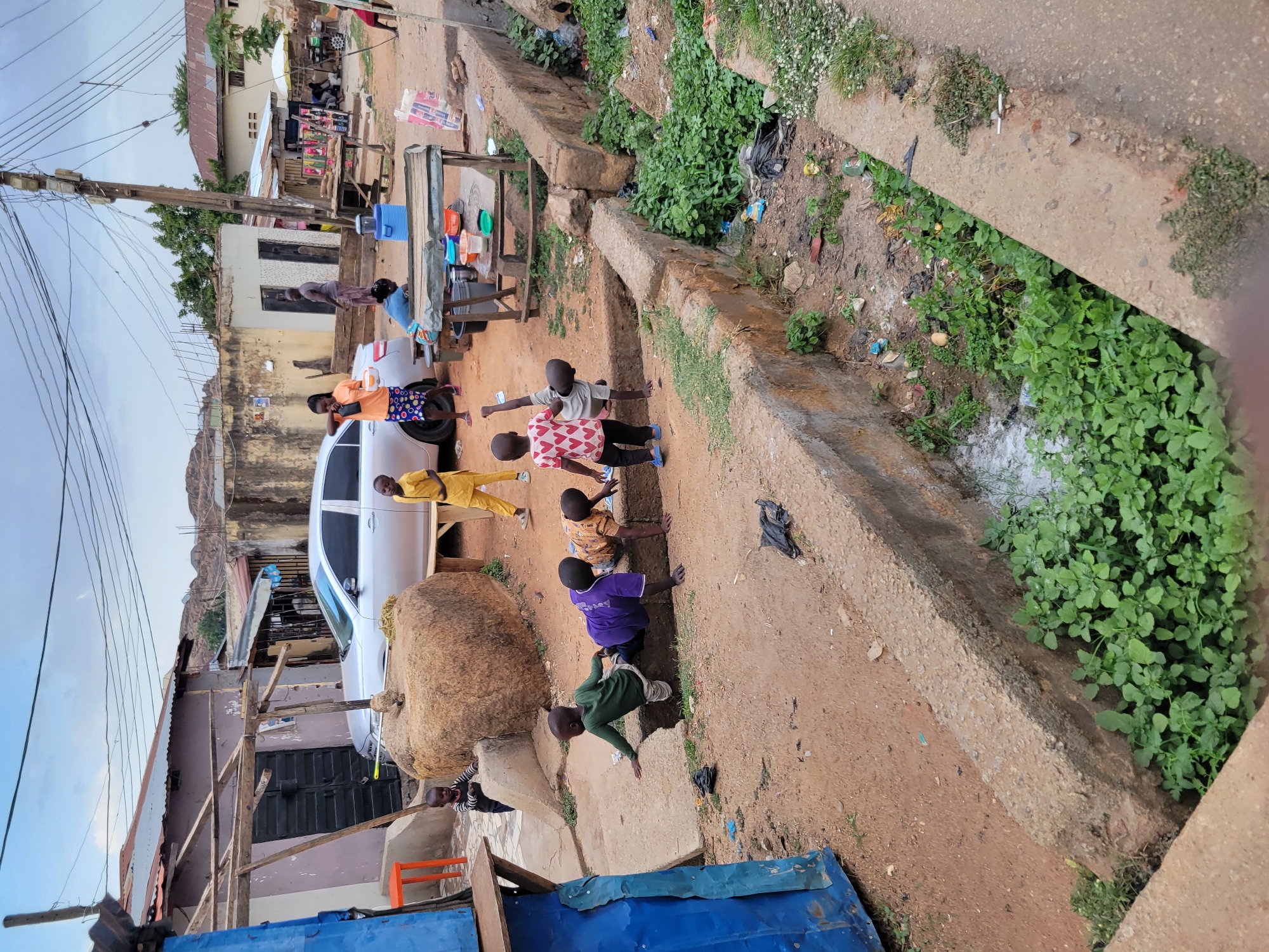 A street scene in Jos, Nigeria, shows several children playing on a dirt road adjacent to a concrete drainage ditch filled with vegetation. A white car is parked nearby. A table with containers sits next to the car, possibly used for selling goods. Buildings and structures are visible in the background, indicating a populated area.