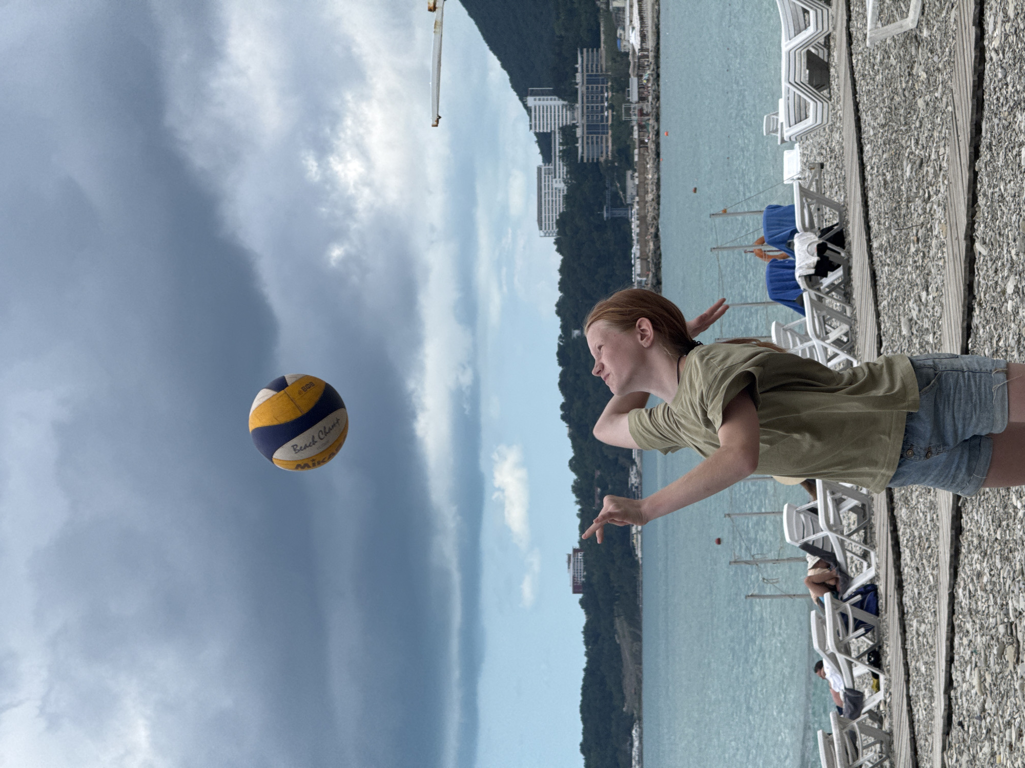 The image depicts a young person with red hair playing volleyball on a beach in Nebug, Russia. The person is wearing a green t-shirt and denim shorts, and is in the process of hitting a volleyball that is suspended in the air above their head. The background shows the turquoise sea, a coastline with buildings, and a cloudy sky. In the foreground, there are rows of empty lounge chairs on a pebble beach, suggesting it is not peak season or time of day. The overall atmosphere is one of leisure and recreation.