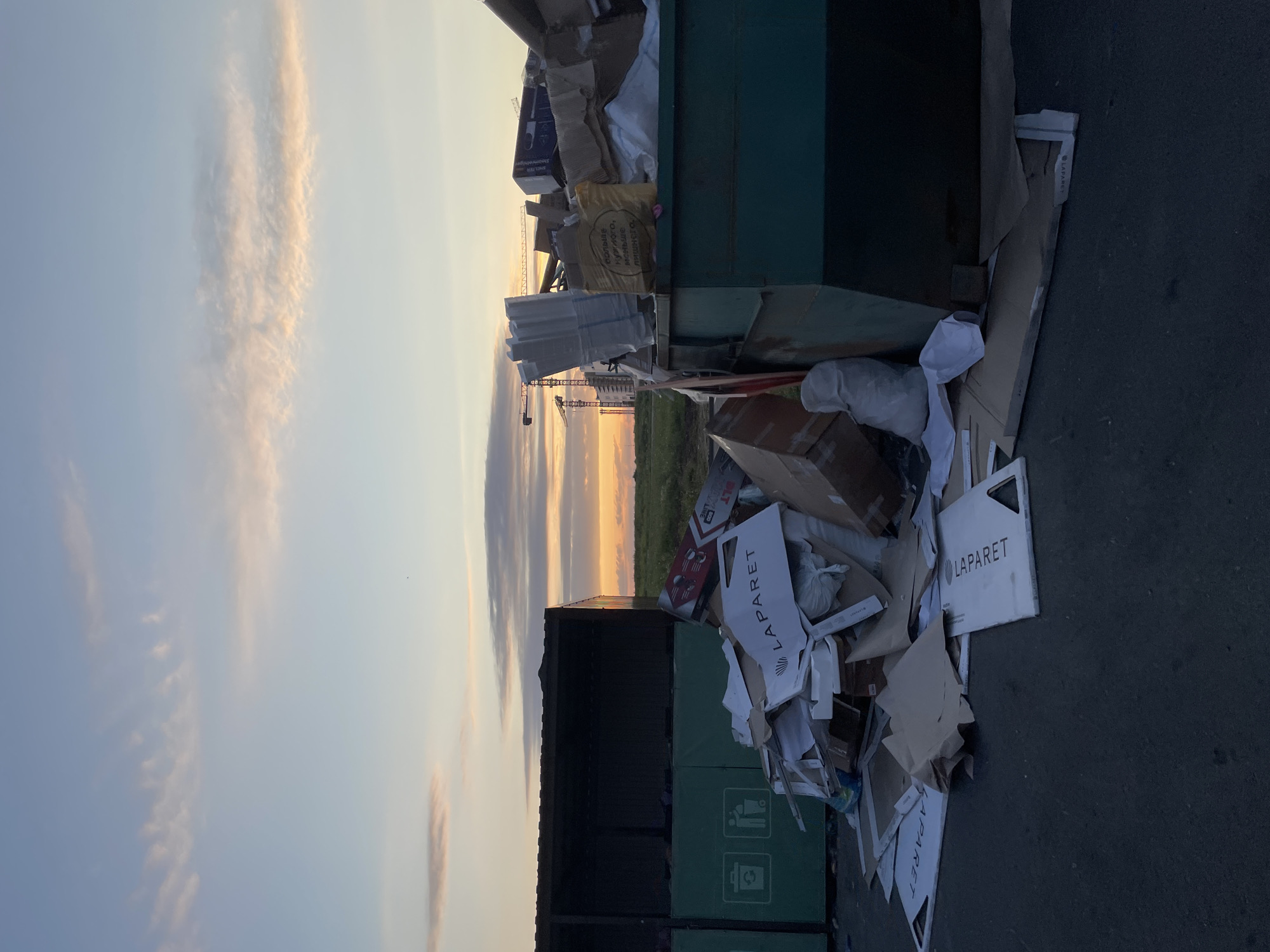 The image depicts an outdoor scene at what appears to be a recycling or waste disposal area, possibly on the outskirts of Stavropol, Russia. The sky, illuminated by the warm hues of a sunset or sunrise, features scattered clouds in shades of pink, orange, and blue. In the foreground, a large green dumpster is overflowing with various types of waste, primarily cardboard boxes, paper packaging, and some plastic. Some of the cardboard pieces display visible text, including the word "LAPARET". To the left, a dark-colored, open-topped structure, possibly another dumpster or storage unit, is visible with green panels bearing recycling symbols. The ground is paved, and in the background, construction cranes and buildings suggest ongoing development in the area. The overall scene is one of urban waste management against a natural, albeit moody, sky. There are no people visible in the image.