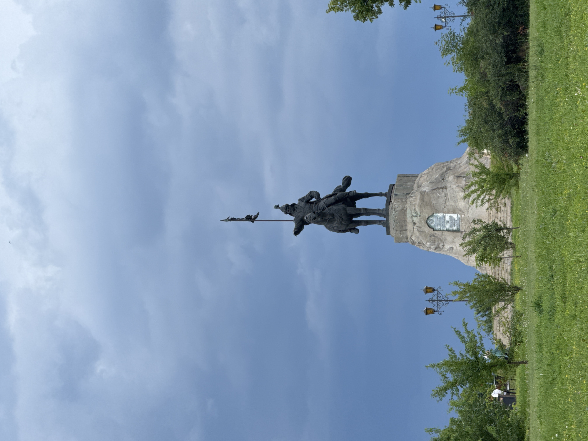 A monumental statue of a warrior on horseback, holding a flag aloft, stands atop a rocky, artificial mound under a dramatic, cloudy sky. The statue is made of dark metal, possibly bronze, and depicts the warrior in historical attire, including a helmet. The mound has a textured, uneven surface, resembling natural stone, with a plaque or inscription visible on its side. In the foreground, a lush green grassy area is dotted with small white flowers and dandelions. Several young trees with vibrant green foliage are planted in a line, leading towards the mound. In the distance, to the left of the statue, two street lamps with ornate, dark metal posts and multiple amber lights are visible. The overall scene suggests an outdoor public space, possibly a park or memorial area.
This statue is located in Yelabuga, Russia, and it is a monument to General Ivan Panfilov, a Soviet military leader who defended Moscow during World War II. The location is identified as Panfilov Park.