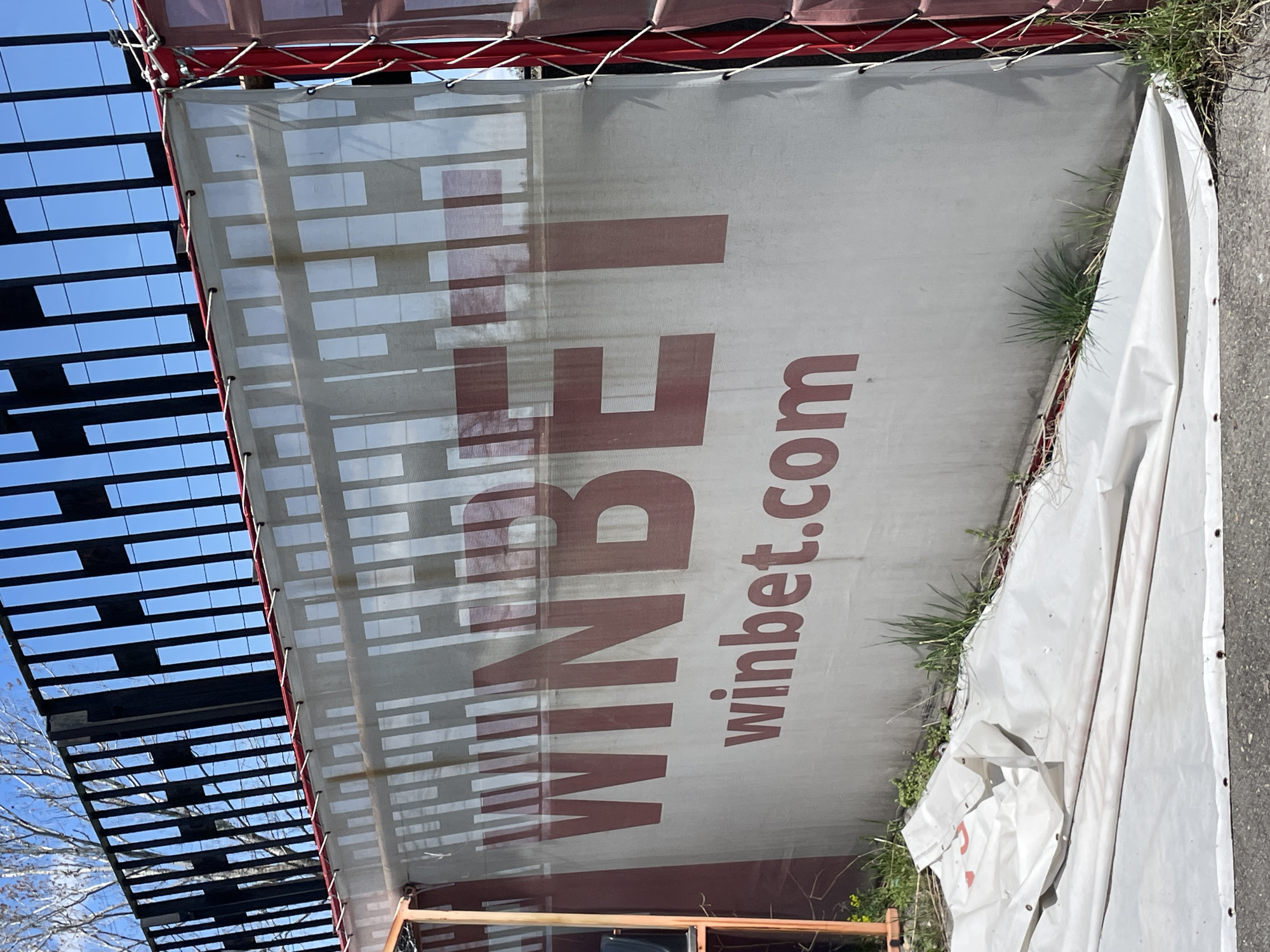 A partially obscured advertisement for "WINBET winbet.com" is attached to a structure with a red frame. The banner itself is white with red text. The advertisement appears slightly faded or weathered. The top of the frame is visible with a black, slatted structure above it, through which the sky is visible. The right edge of the advertisement is frayed, revealing another white section of a similar banner, indicating that this section has been pulled back. Grass and weeds are growing at the bottom edge of the banner. The location is implied to be Sofia, Bulgaria, from the brand associated with the advertisement. The advertisement is outdoors and appears to be situated adjacent to a walkway/paved area.