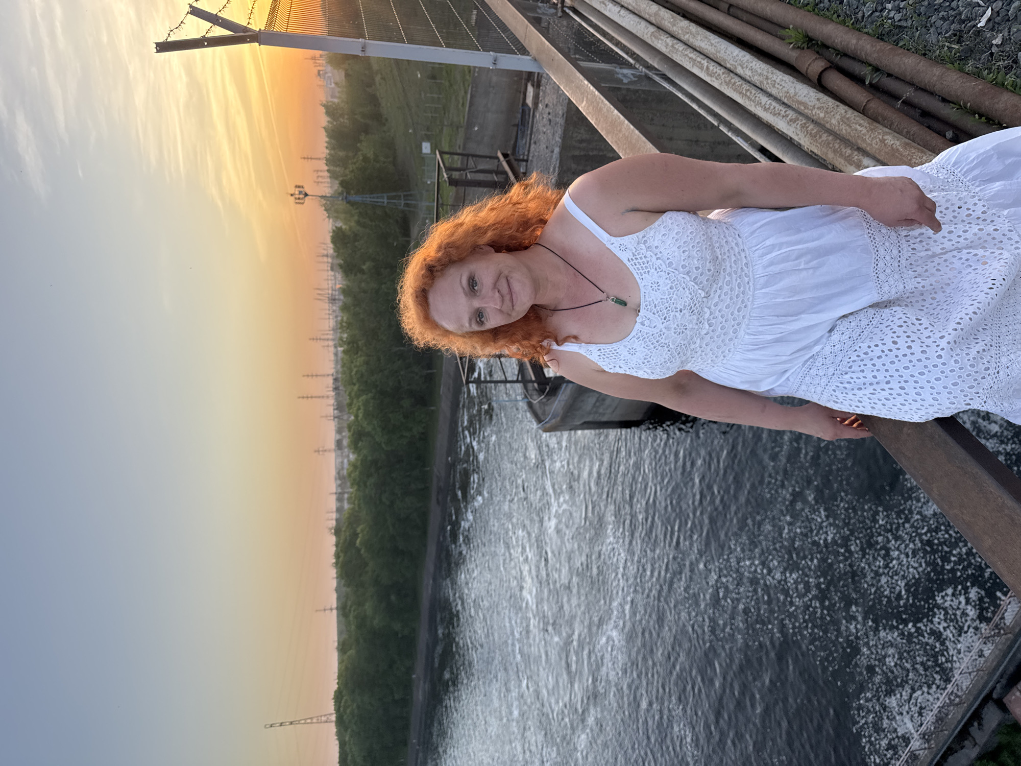 A woman with red curly hair is standing on a metal bridge overlooking a body of water. She is wearing a white, eyelet sundress and a necklace with a green stone. The sun is setting in the background, casting a warm glow on the scene. In the distance are electrical towers and trees. Metal pipes and a chain-link fence are visible along the bridge. This photo was taken from the observation deck located on the Dam-Bridge-Road, which crosses the Kama River in Chaikovsky, Russia.