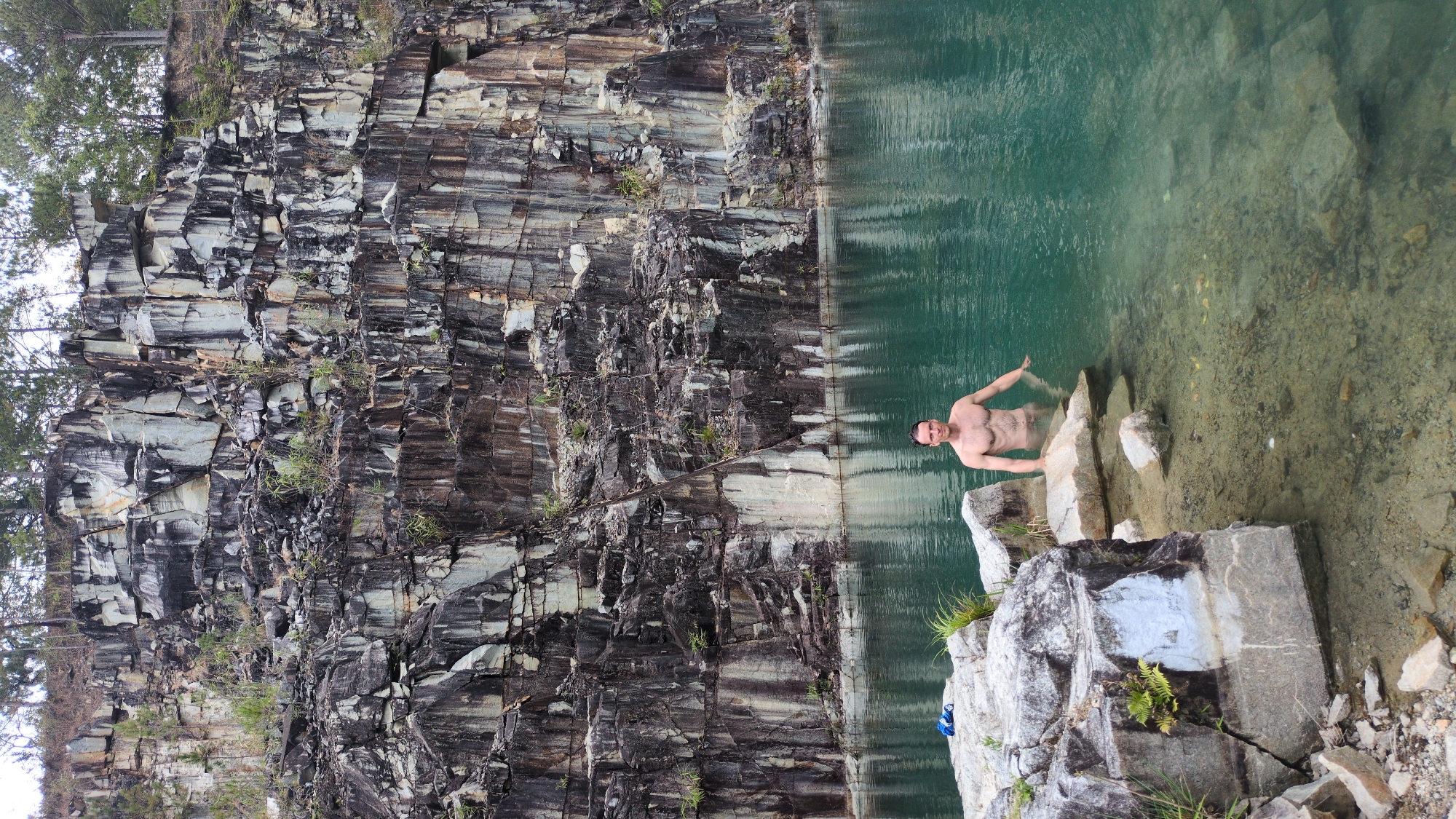 A vertically oriented medium shot depicts a nude man in a body of water next to a rocky cliff.
The cliff face, composed of dark gray and beige striated stone, takes up the left half of the image. Small green plants grow intermittently on its surface. The water, a translucent green hue, occupies the right half. The man is waist-deep in the water, standing on rocks. He has short dark hair and fair skin. His arms are partially submerged, extended to the sides. His expression is neutral. He is positioned between the main body of water and a collection of larger rocks at the water's edge. Some vegetation is present on these rocks.
The scene is set at the location in the city of Lang Biang - Da Lat, Vietnam.