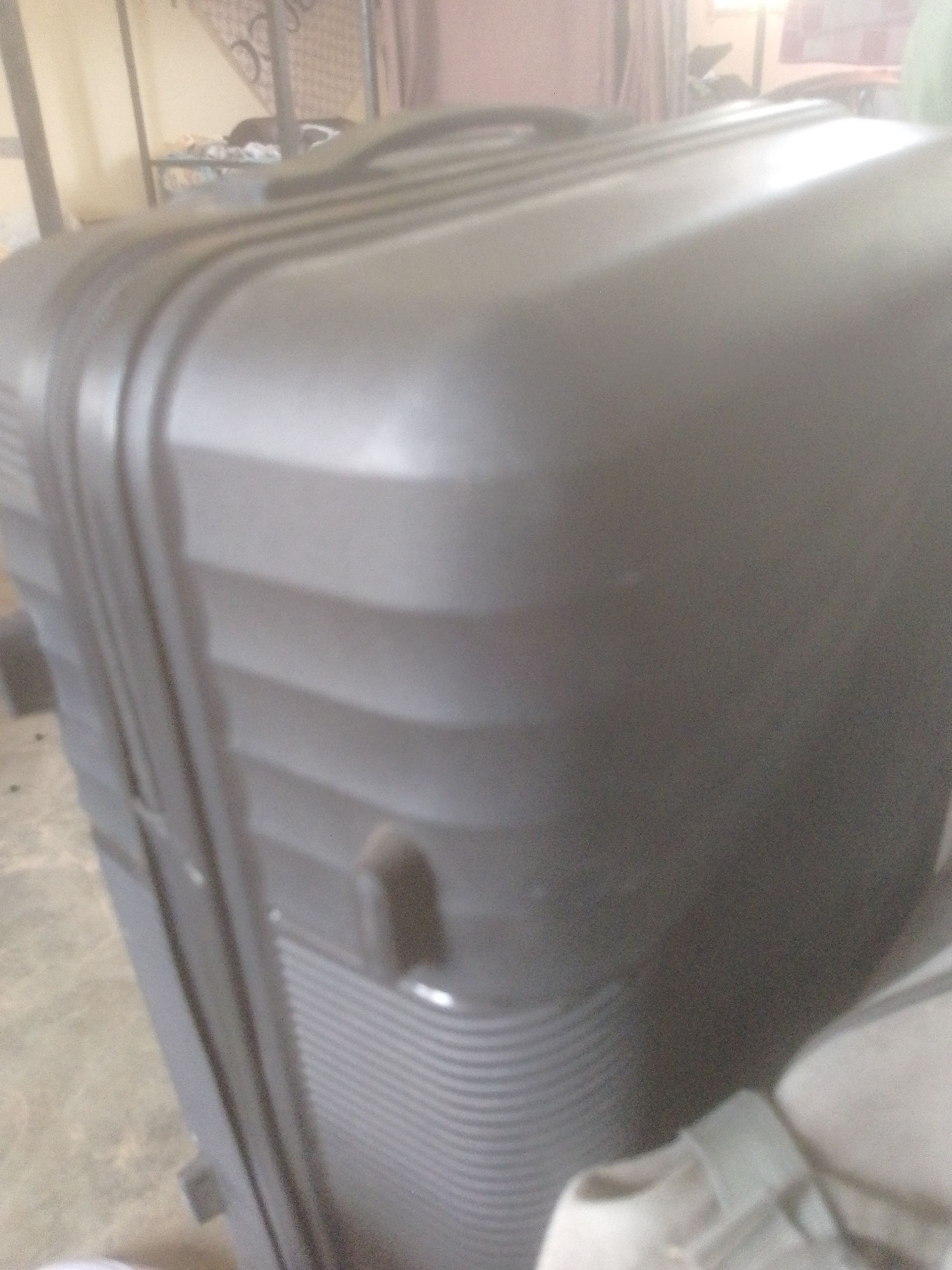 The image shows a close-up of a large, dark gray suitcase in an indoor setting. The suitcase has a textured, ribbed surface. A strap or band runs vertically along the side of the suitcase. The background is blurry and appears to be a room with some furniture, possibly a bed frame and shelving, suggesting it might be a bedroom or storage area. There is a general sense of a domestic or personal space. The location in Danbazau, Nigeria is not directly discernible from the visual information in the image itself.