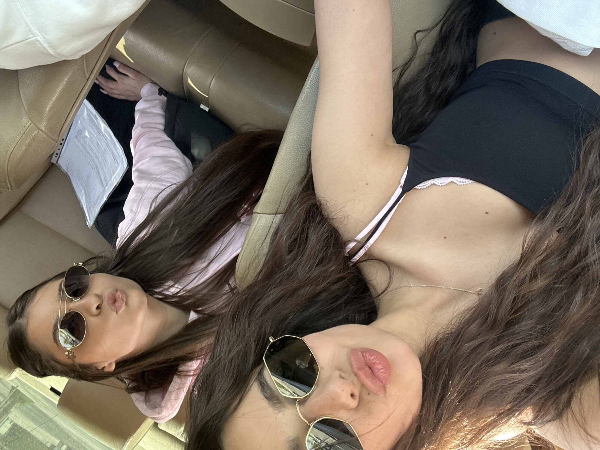 This image captures two young women inside a car, likely in Plovdiv, Bulgaria, during the daytime under bright, sunny conditions. The photo is taken from an overhead perspective, possibly a selfie.
In the foreground, a woman with long, wavy dark brown hair lies with her head tilted, wearing a black sleeveless top with white and pink lace trim and thin straps. She has gold-rimmed octagonal sunglasses on and her lips are slightly pursed, showcasing light pink lipstick. A delicate gold chain necklace is visible around her neck.
Further back, seated in what appears to be the front passenger seat, is another woman with similar long, dark brown hair. She is wearing a pink long-sleeved top or hoodie and round, gold-rimmed sunglasses. Her lips are also pursed in a playful "duck face" pose.
The car's interior features light beige or tan leather seats, and part of a car armrest is visible. Between the two women, on the passenger seat, a stack of white papers with illegible text is present. Both women appear relaxed and are posing playfully for the camera.