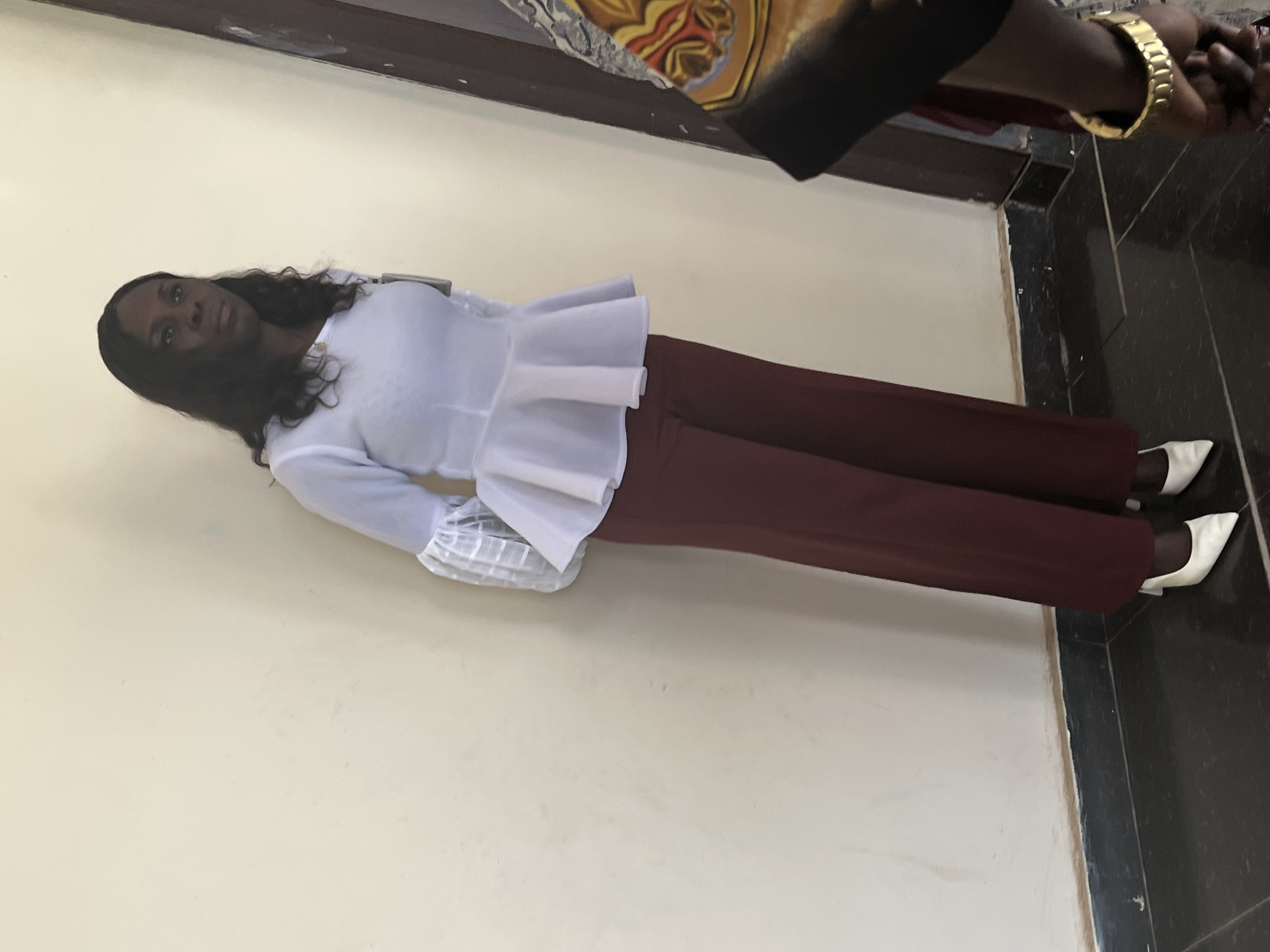 A woman with dark skin and long black hair is standing against a light-colored wall. She is wearing a white blouse with a ruffled hem and long sleeves, paired with maroon-colored pants and white pointed heels. The location appears to be indoors, possibly in a building in Abuja, Nigeria. The floor adjacent to the wall is covered in dark gray tiles. A person wearing a gold watch is partially visible in the upper-right corner of the image.
